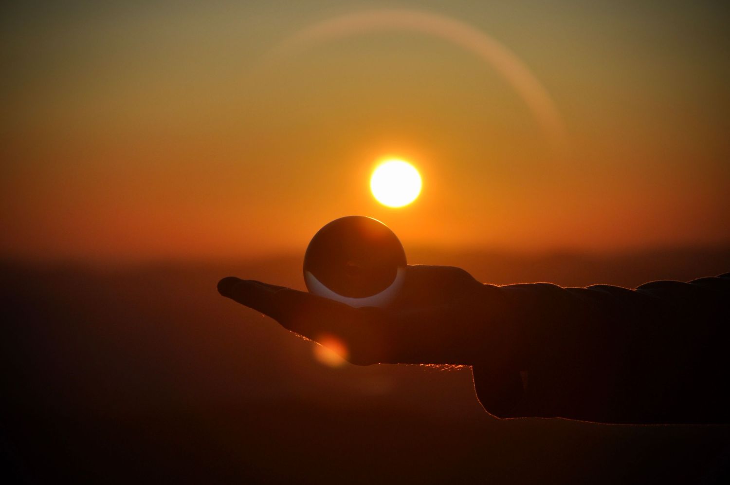 Silhouetted hand balancing a glass sphere against a vibrant orange sunset sky.
