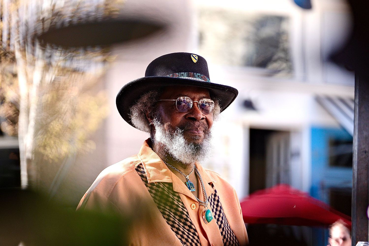 Stylish older man with white beard wearing a wide-brim hat outdoors in sunlight.