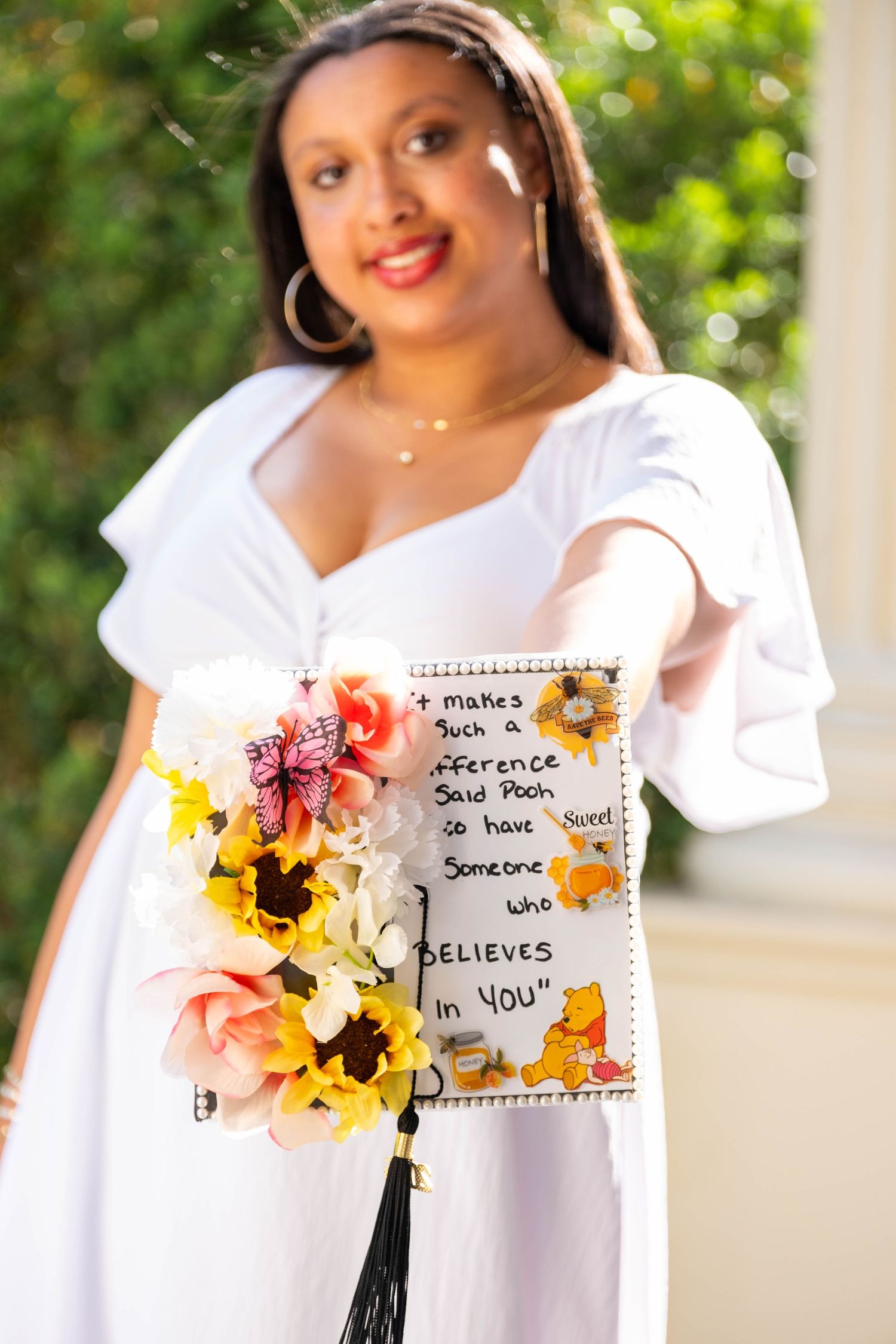 Graduate in white dress holds decorated cap with Winnie the Pooh quote and colorful flower bouquet outdoors.