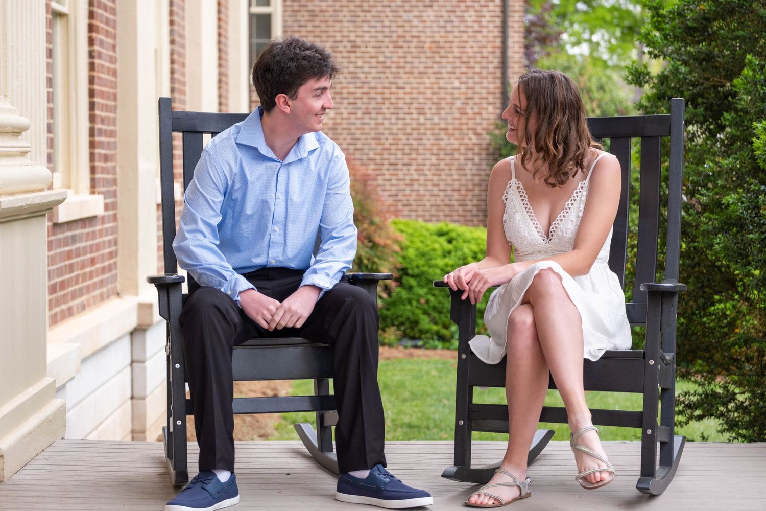 Young couple sitting in black rocking chairs on a porch, smiling at each other during a casual outdoor photo session.