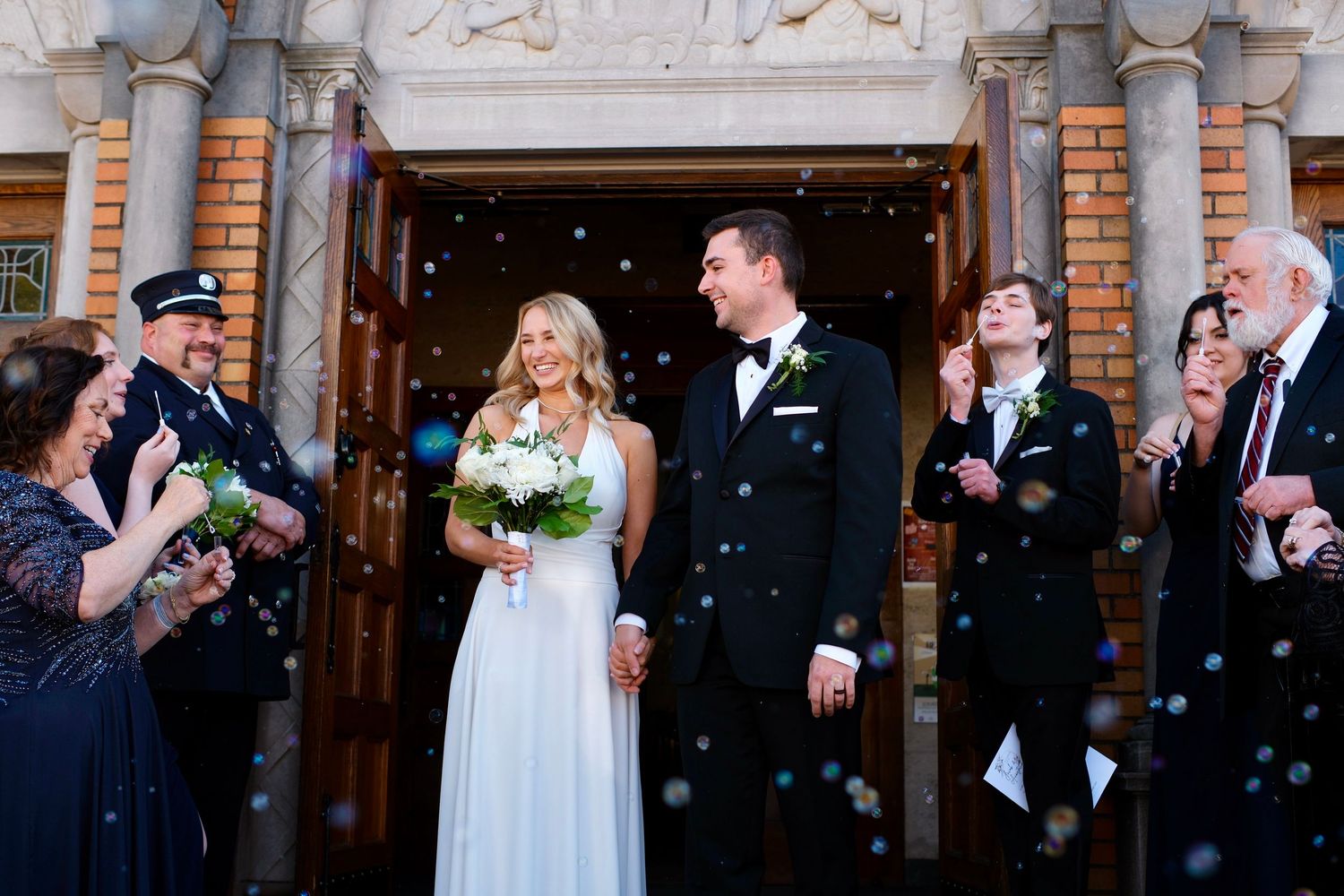 Newlyweds exit church doors as guests celebrate with bubbles after wedding ceremony.