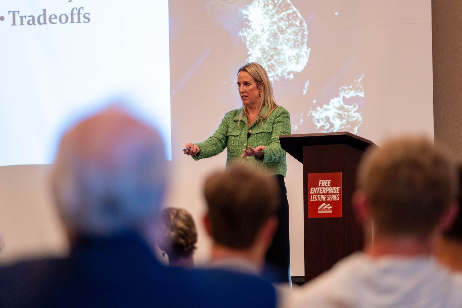 Woman in green jacket presenting at Free Enterprise Lecture Series podium, discussing tradeoffs to seated audience.