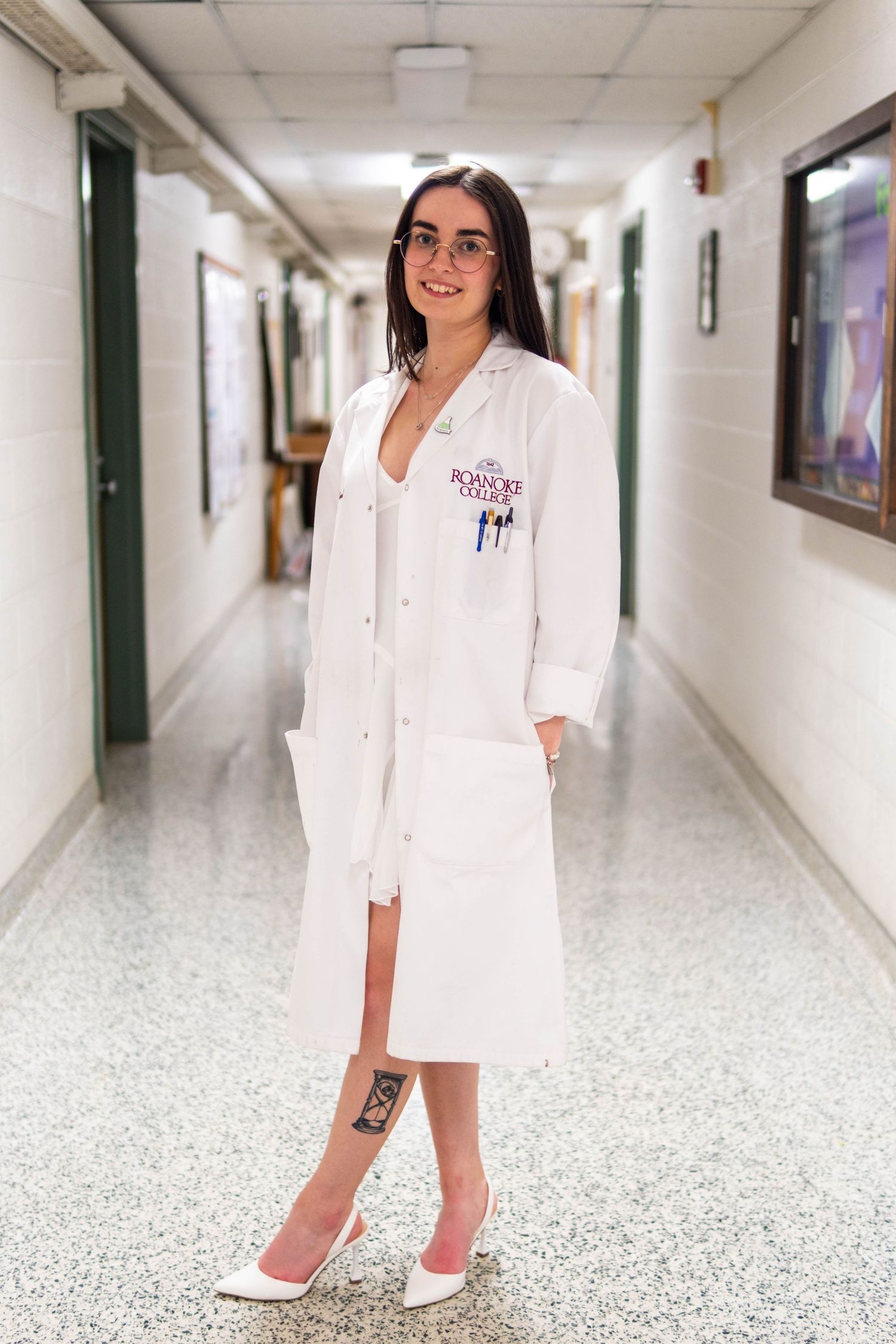 Woman in white lab coat and heels smiling in a hospital hallway, wearing glasses and a Roanoke College emblem.
