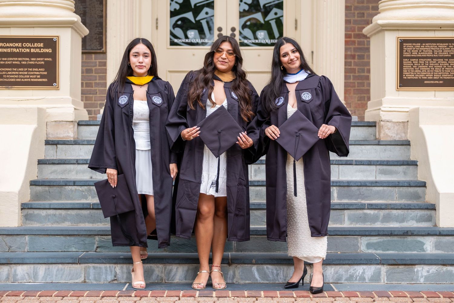 Three women in graduation gowns pose proudly on brick steps outside a college administration building.