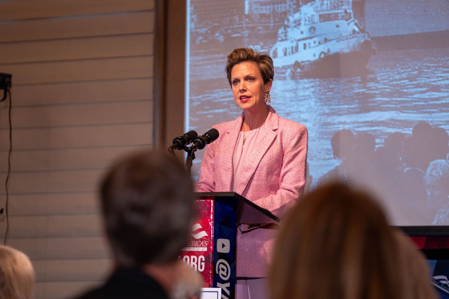 Blonde woman in pink blazer speaking at podium during indoor conference event.