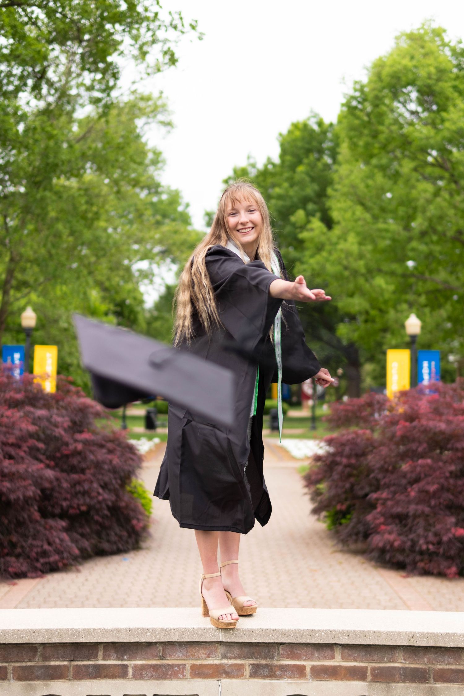 Happy female graduate tossing her cap while wearing black gown on a tree-lined campus walkway.