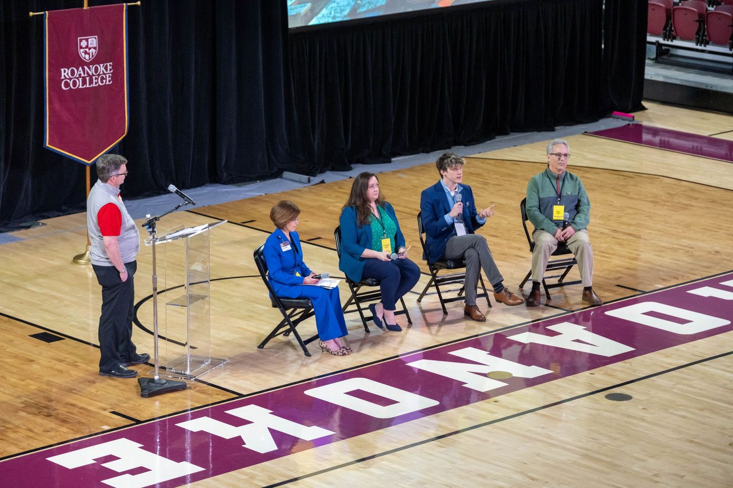 Panel discussion on Roanoke College gymnasium floor with four seated panelists and one student at podium.