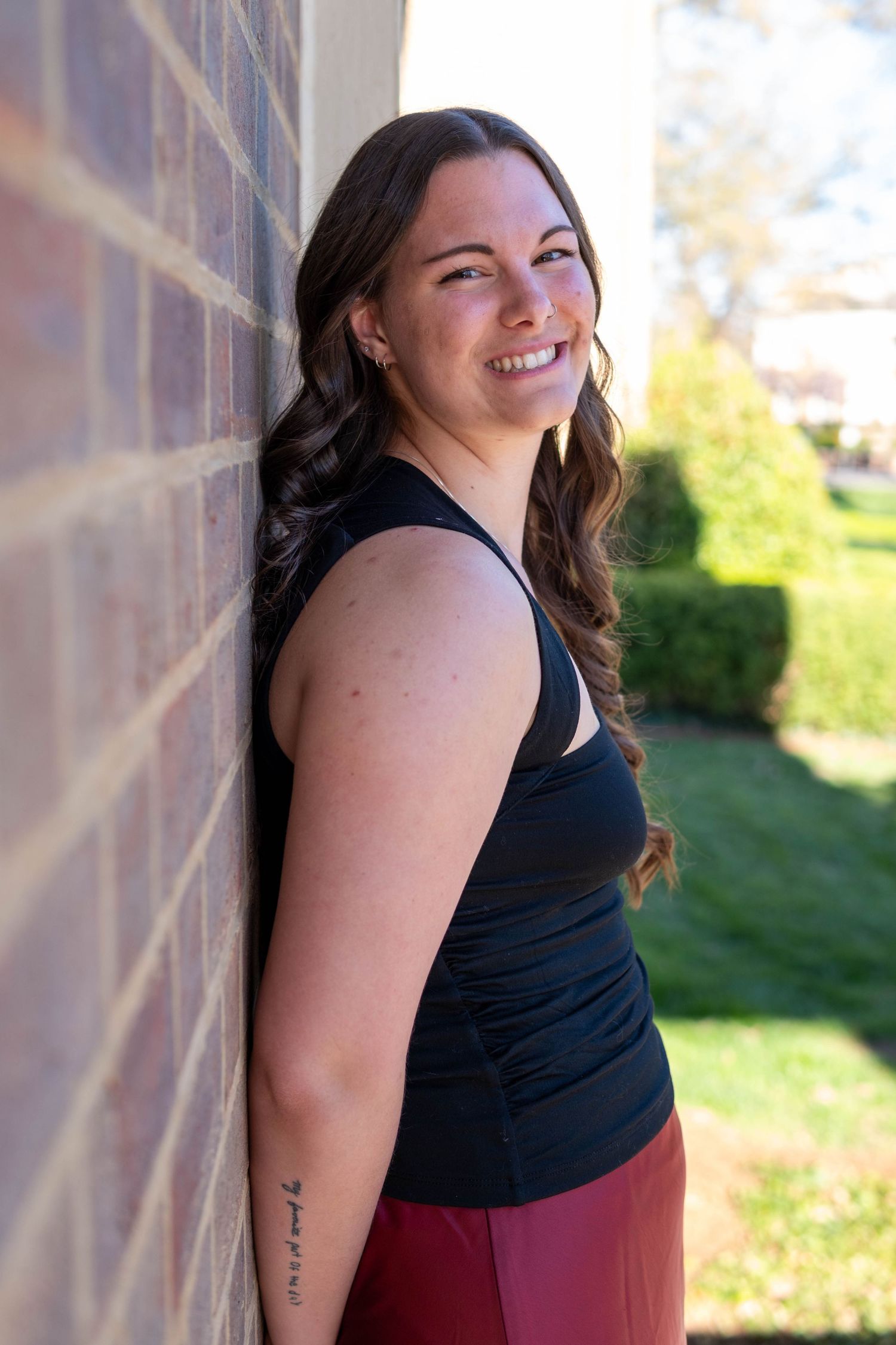 Smiling young woman with wavy brown hair leaning against brick wall in black tank top outdoors.