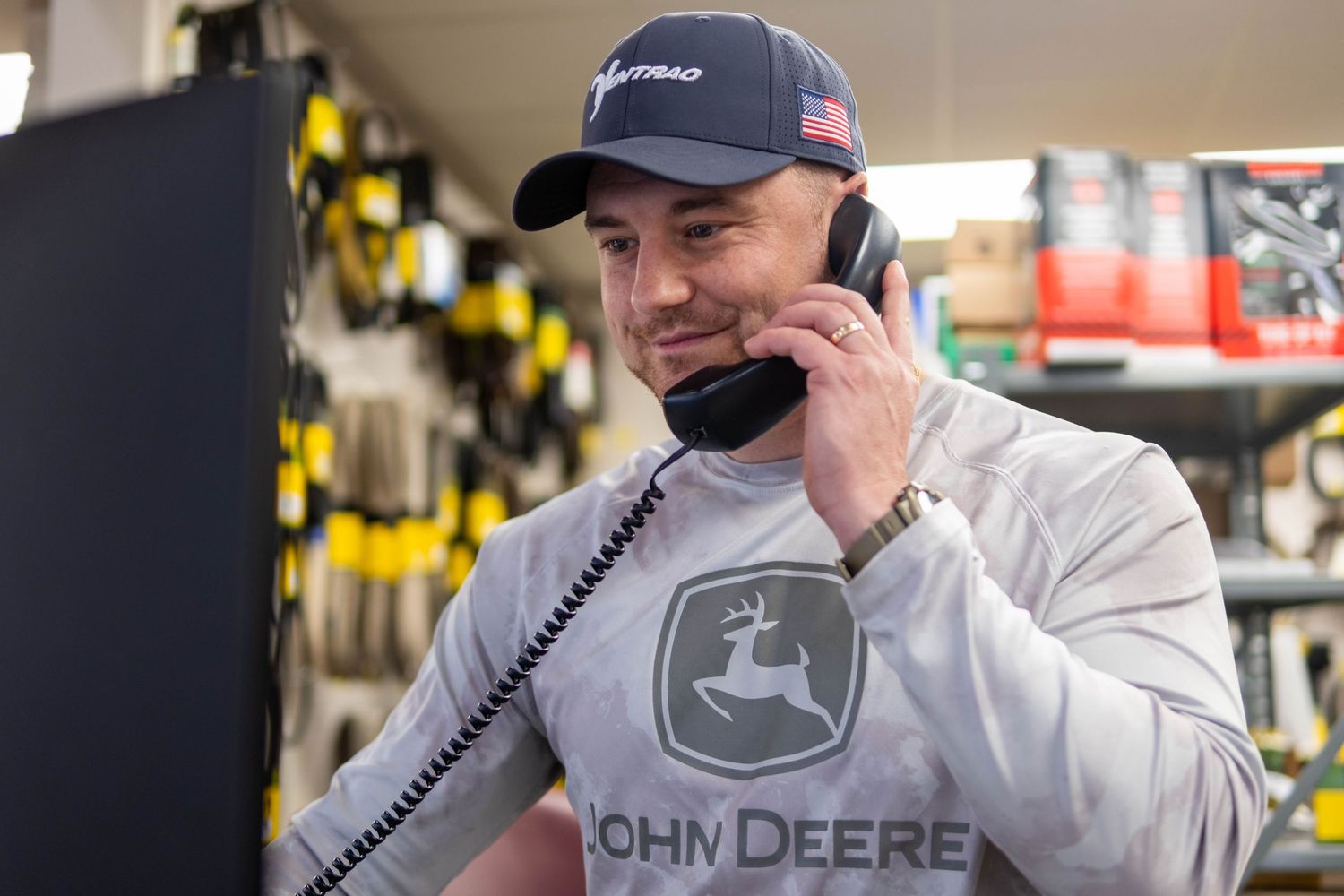 A man wearing a John Deere shirt and cap talks on a corded phone in a parts store.