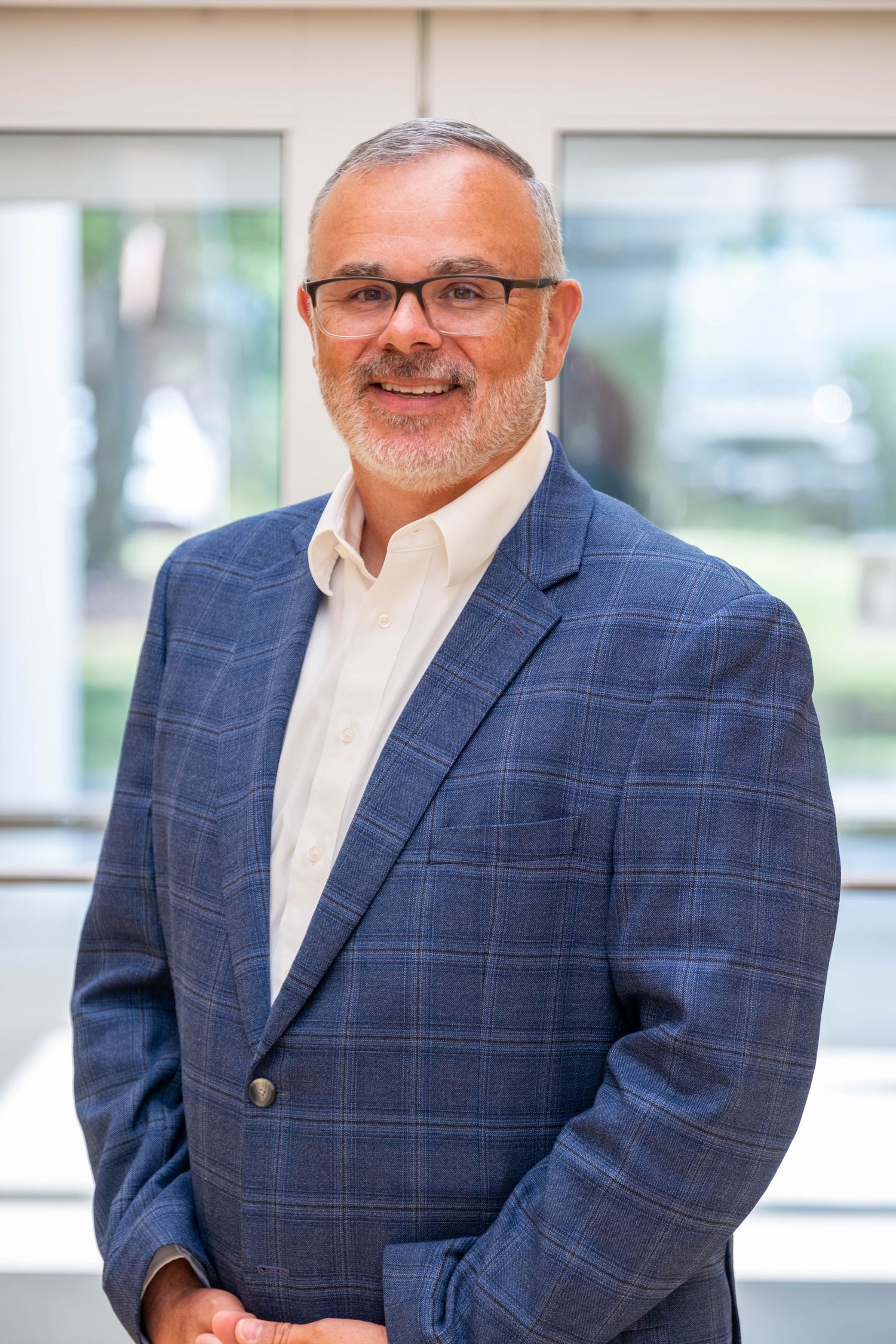 Middle-aged man with glasses and gray beard wearing a blue plaid blazer smiling in a bright professional office setting.
