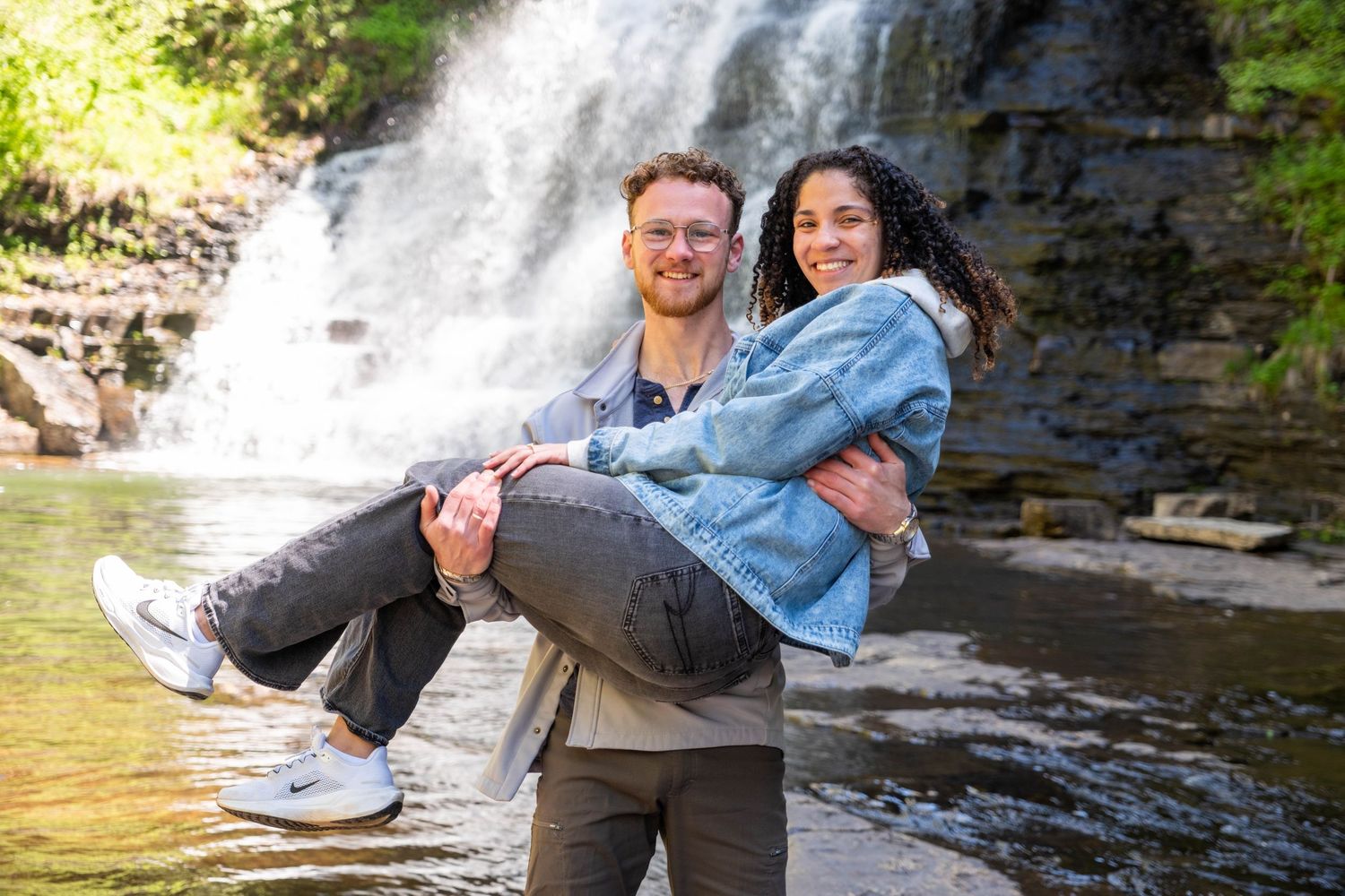 Couple posing by a waterfall, man carrying woman in his arms, smiling outdoors in nature.
