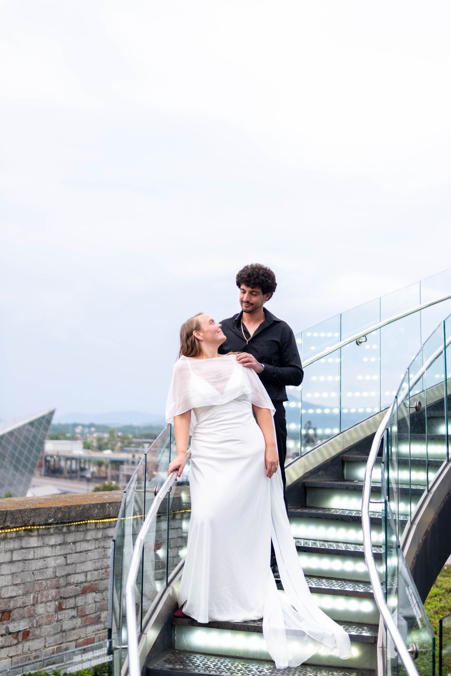 Bride in white gown and groom in black shirt share a romantic moment on outdoor stairs with city views.