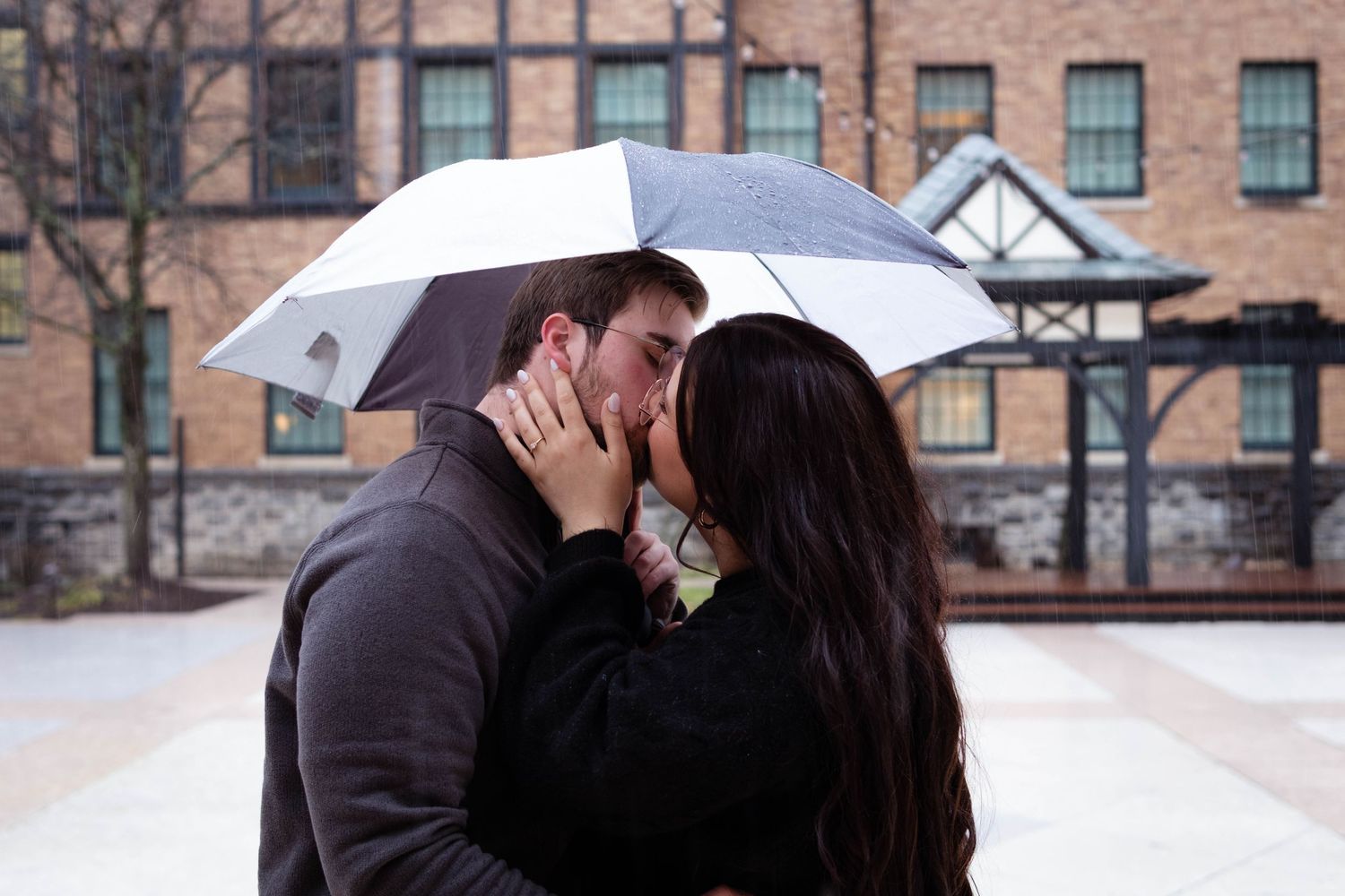 Romantic couple kissing under white umbrella in rainy courtyard with brick building in background.