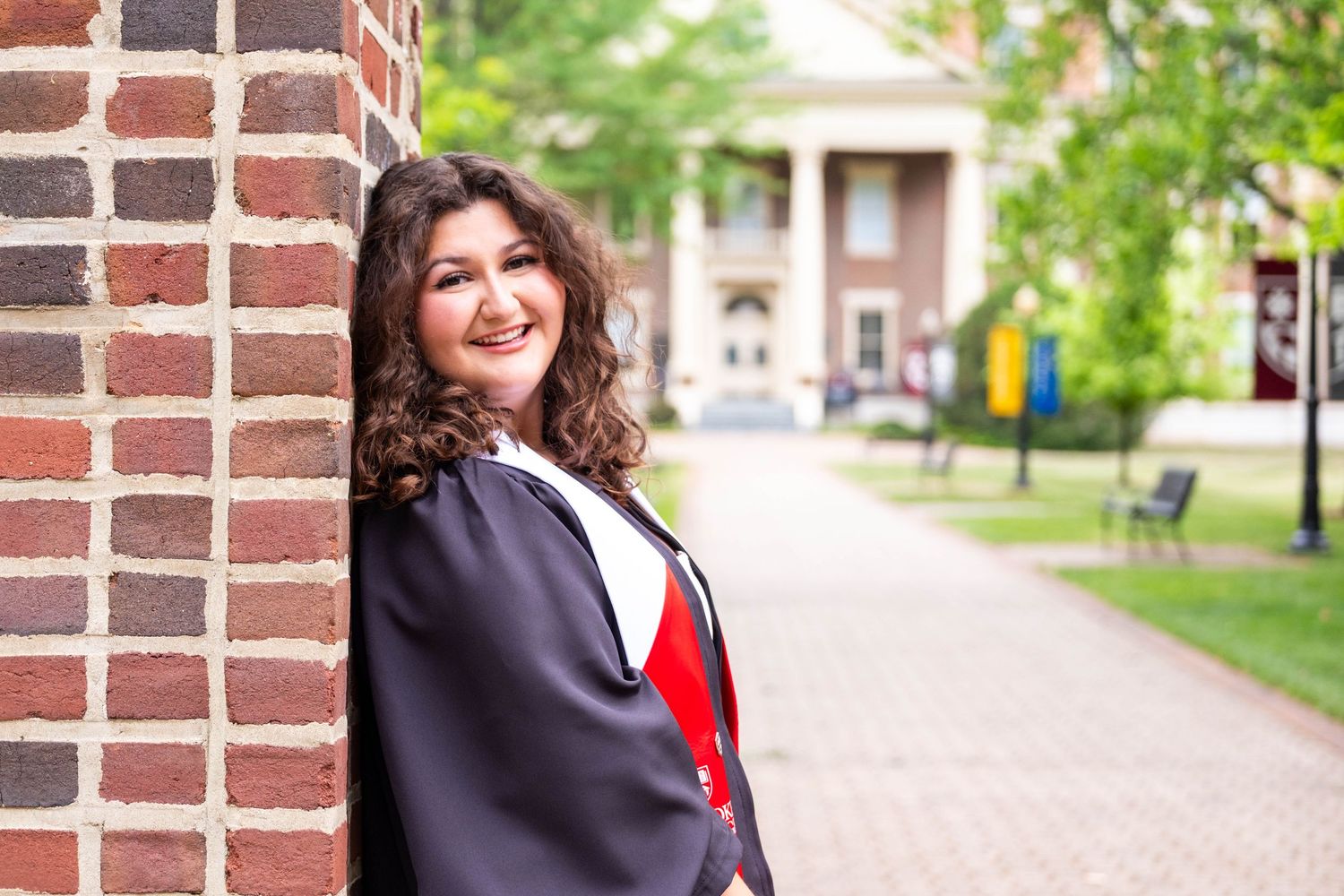 Young woman in graduation gown leaning against brick wall on college campus, smiling proudly on graduation day.