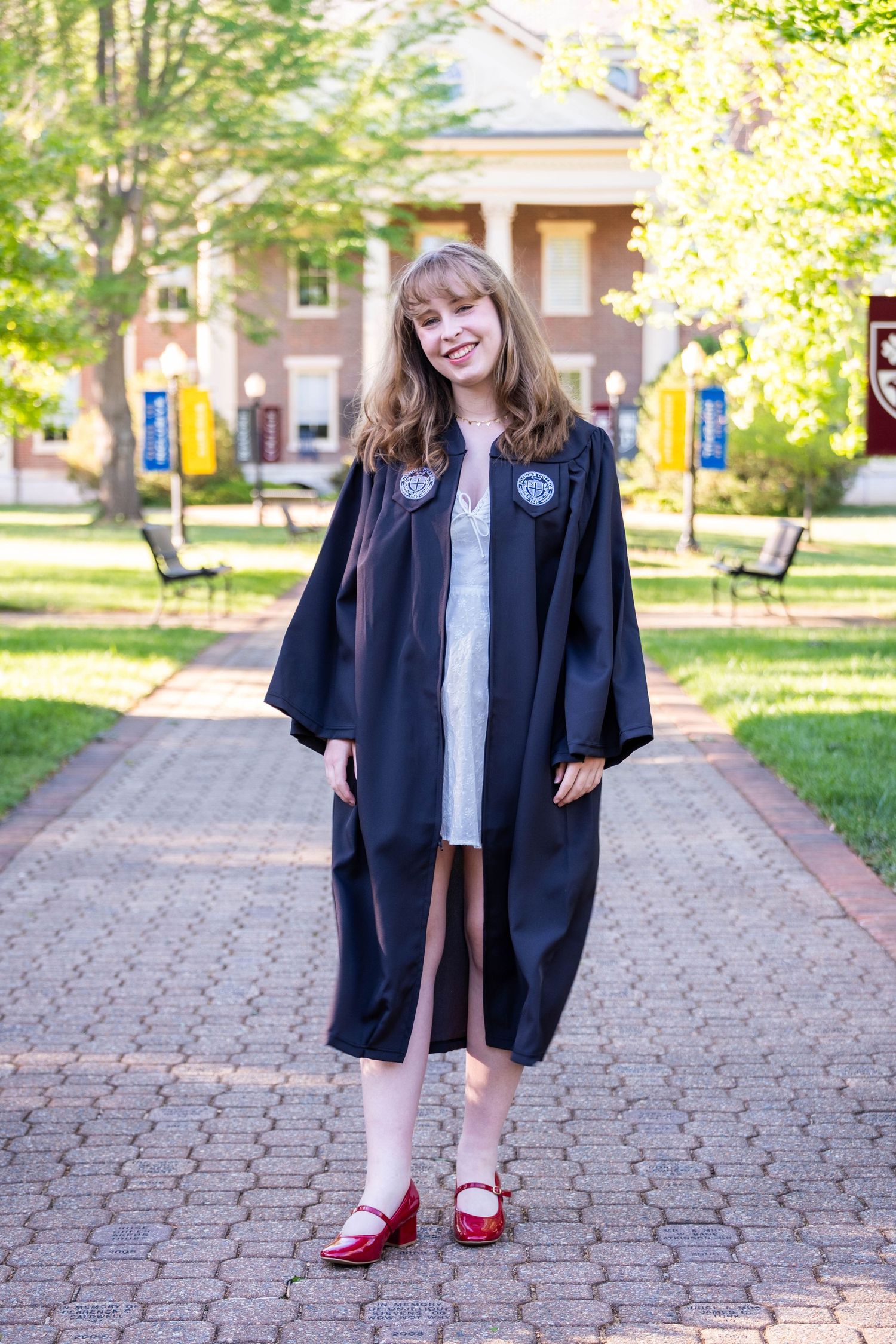 Young woman in black graduation gown and red shoes standing on brick path outside a college building.