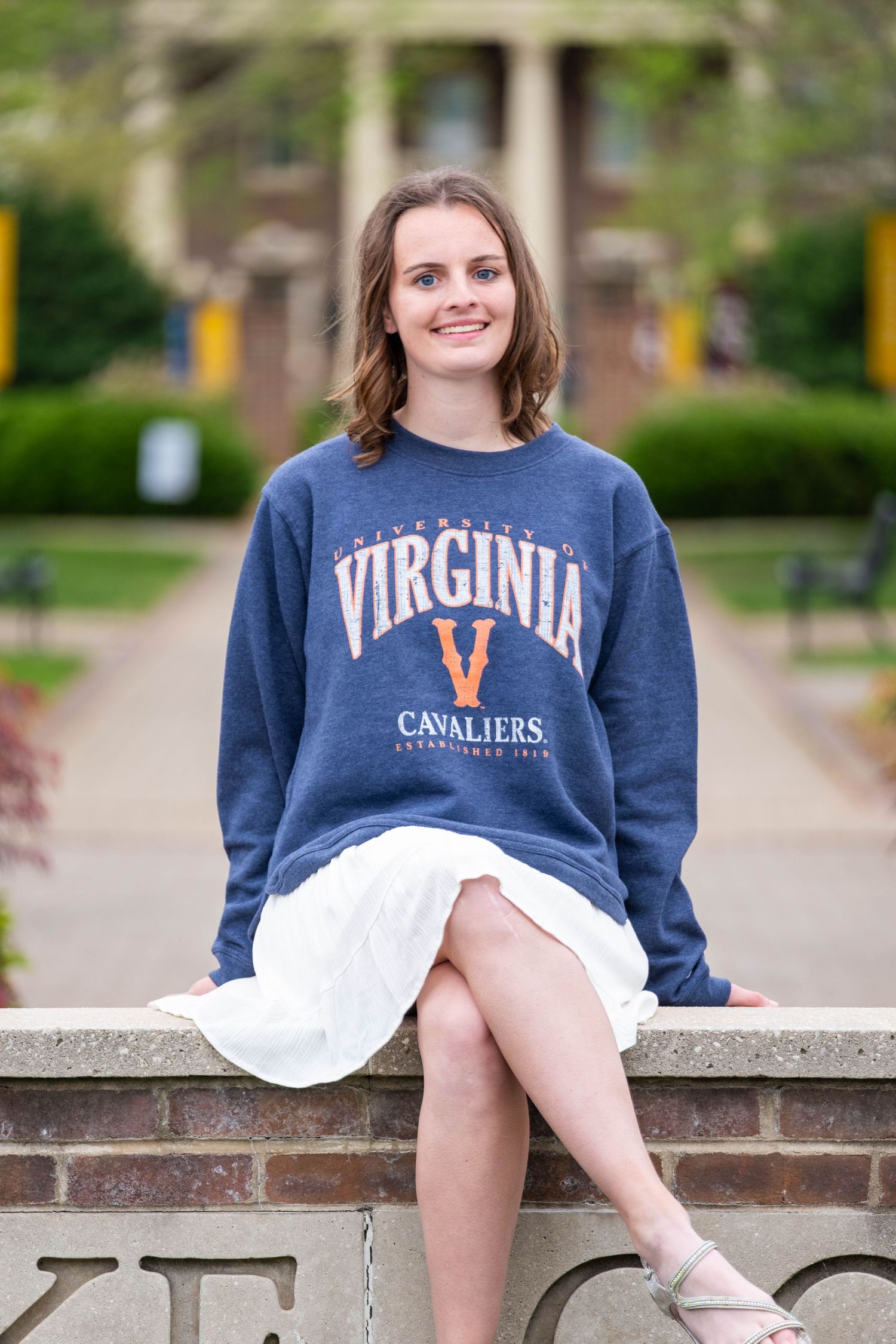 Young woman in University of Virginia Cavaliers sweatshirt sitting on brick wall outdoors on campus.