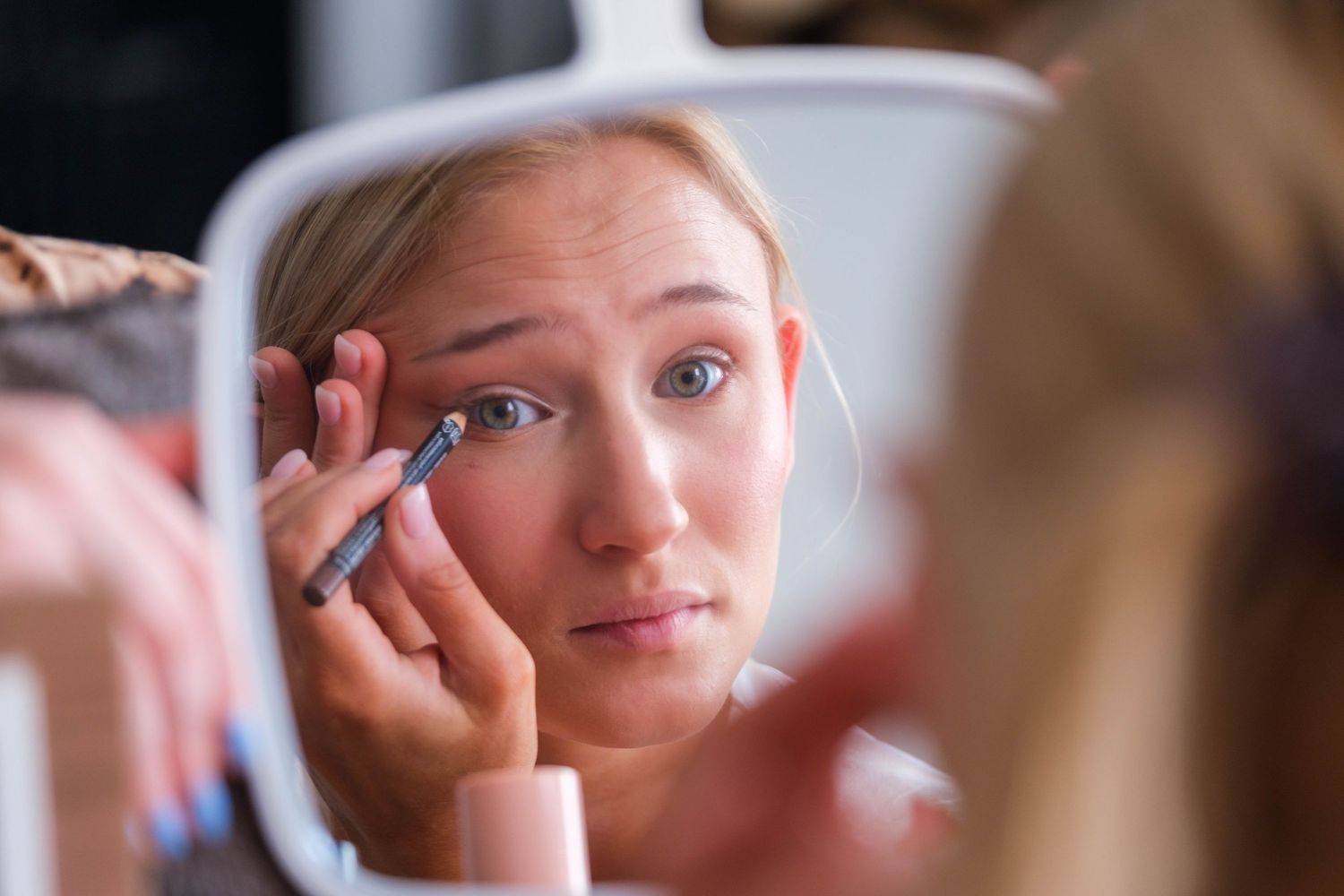 Young blonde woman applying eyeliner while looking in a handheld mirror during makeup routine.