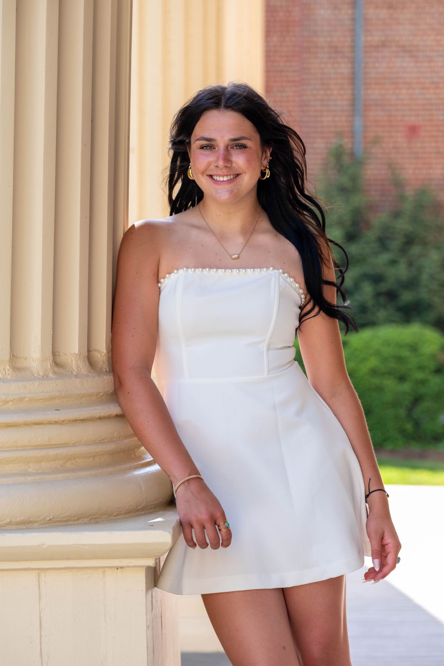 Young woman in white strapless mini dress smiling beside a classical column outdoors.