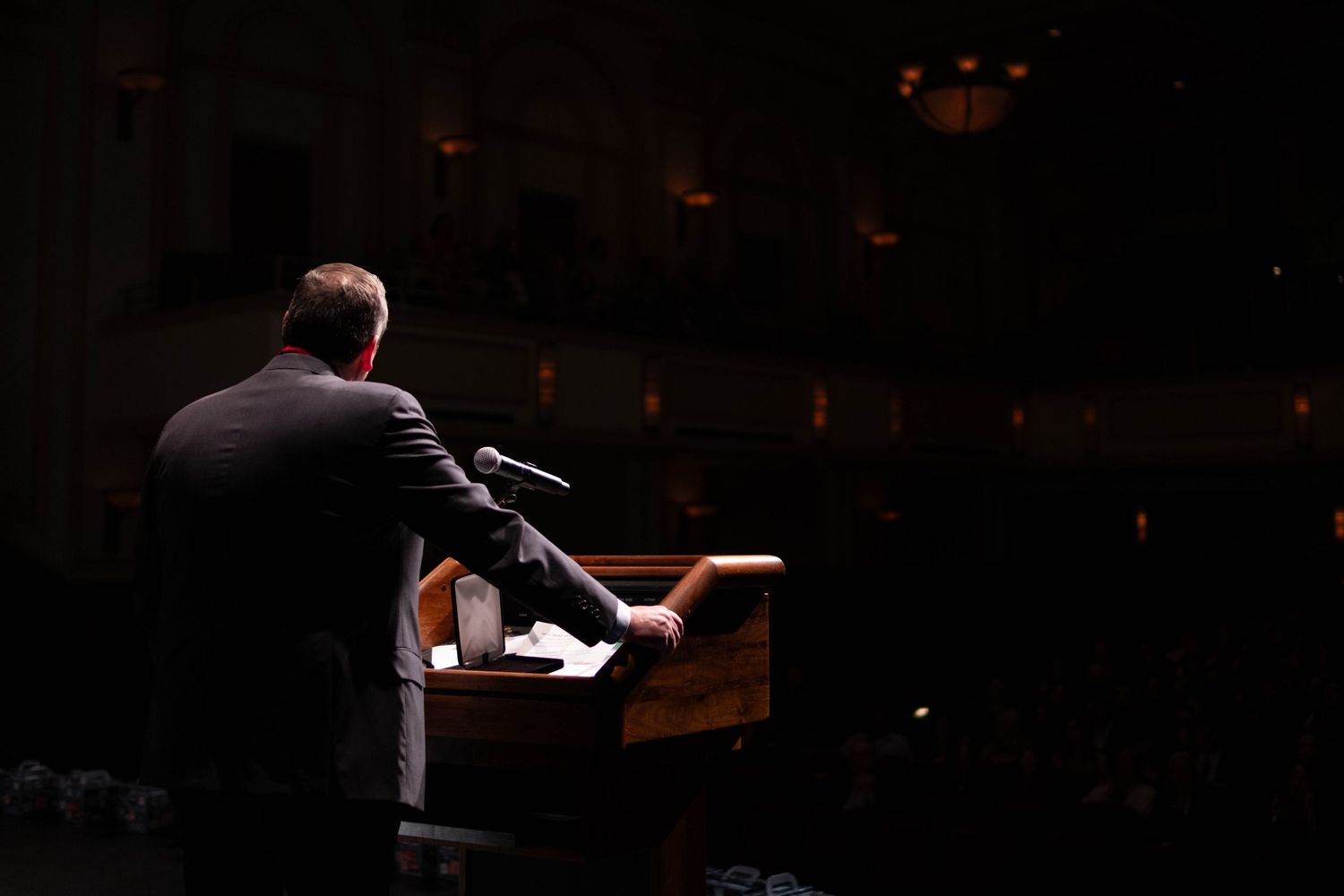 Speaker stands at illuminated podium addressing large audience in darkened theater venue.