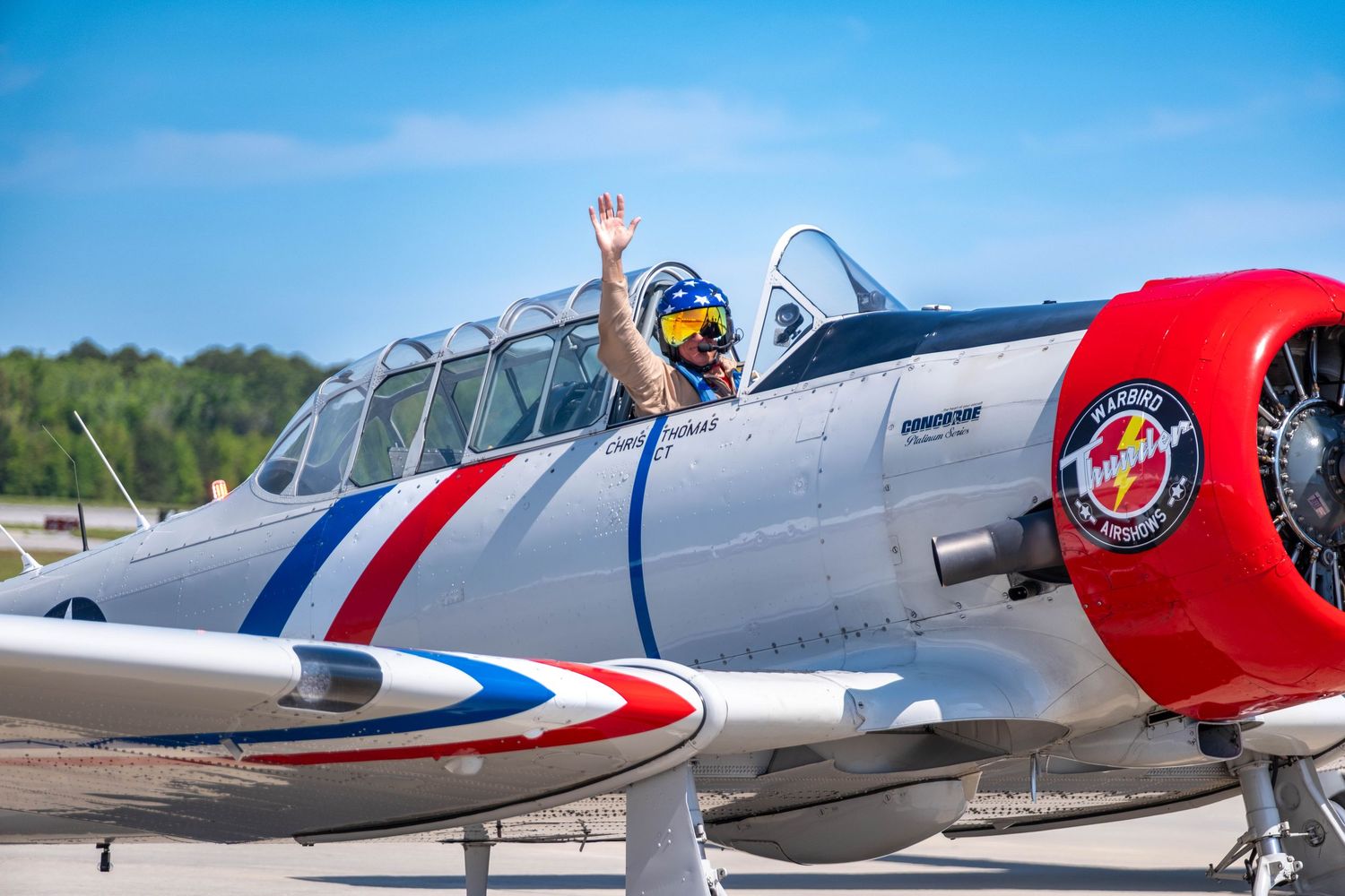 Pilot waving from a red and silver vintage Harvard trainer aircraft at an airshow.