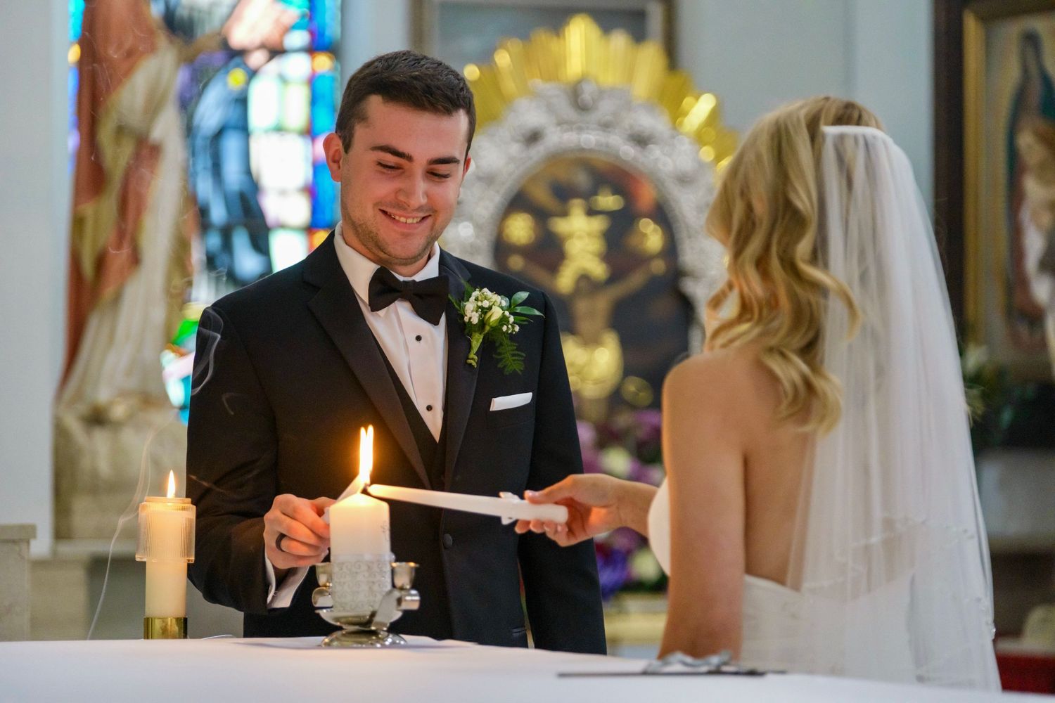Groom in a tuxedo smiling while lighting a unity candle alongside his veiled bride at the altar.