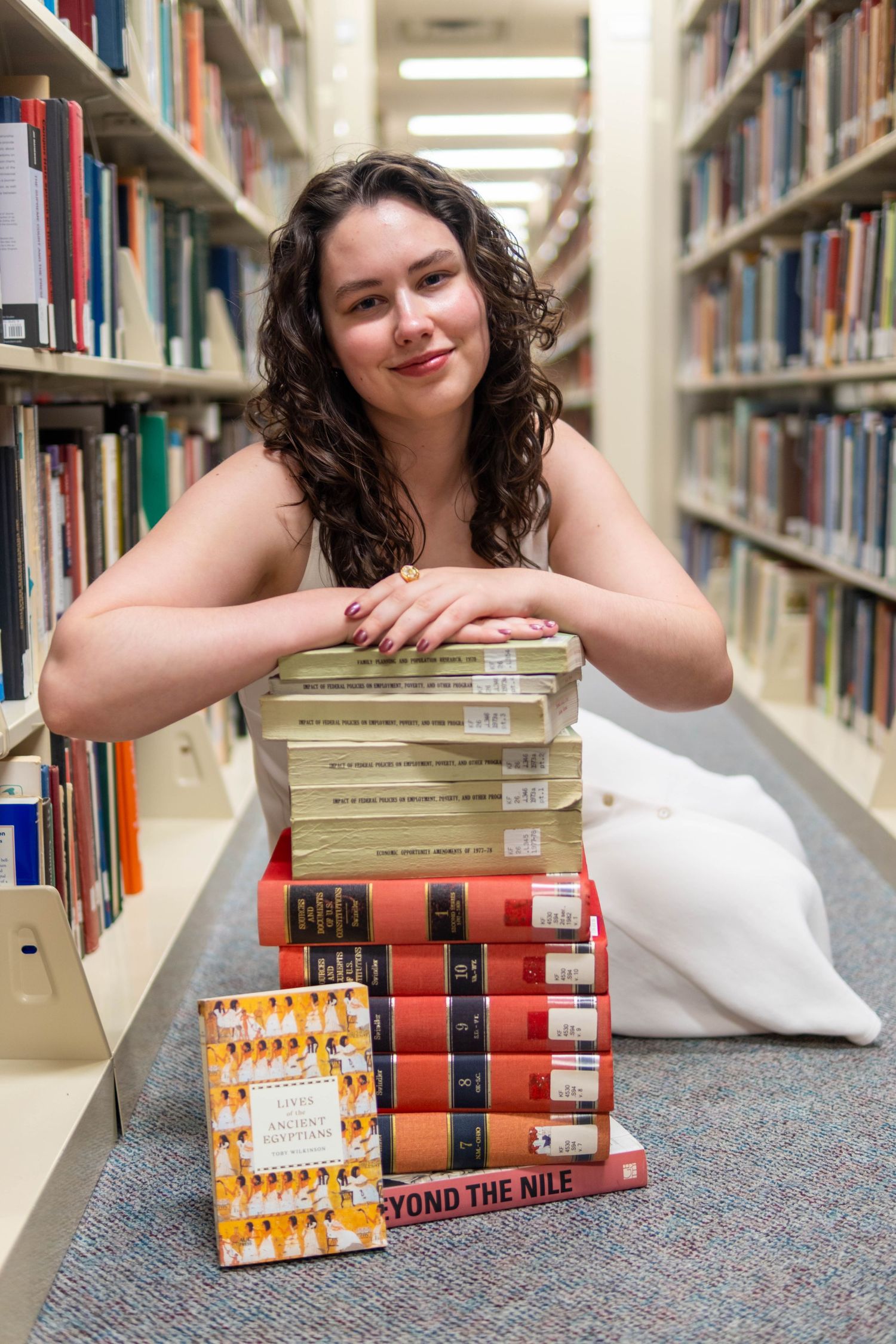 Young woman poses with stacked library books in aisle, including 