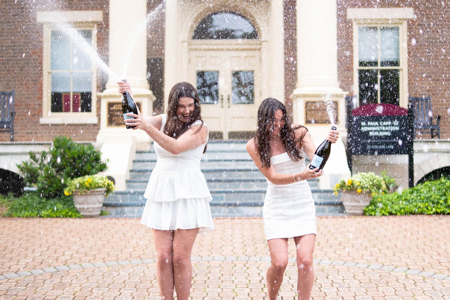 Two women in white dresses celebrating graduation by popping champagne bottles outside a brick university building.