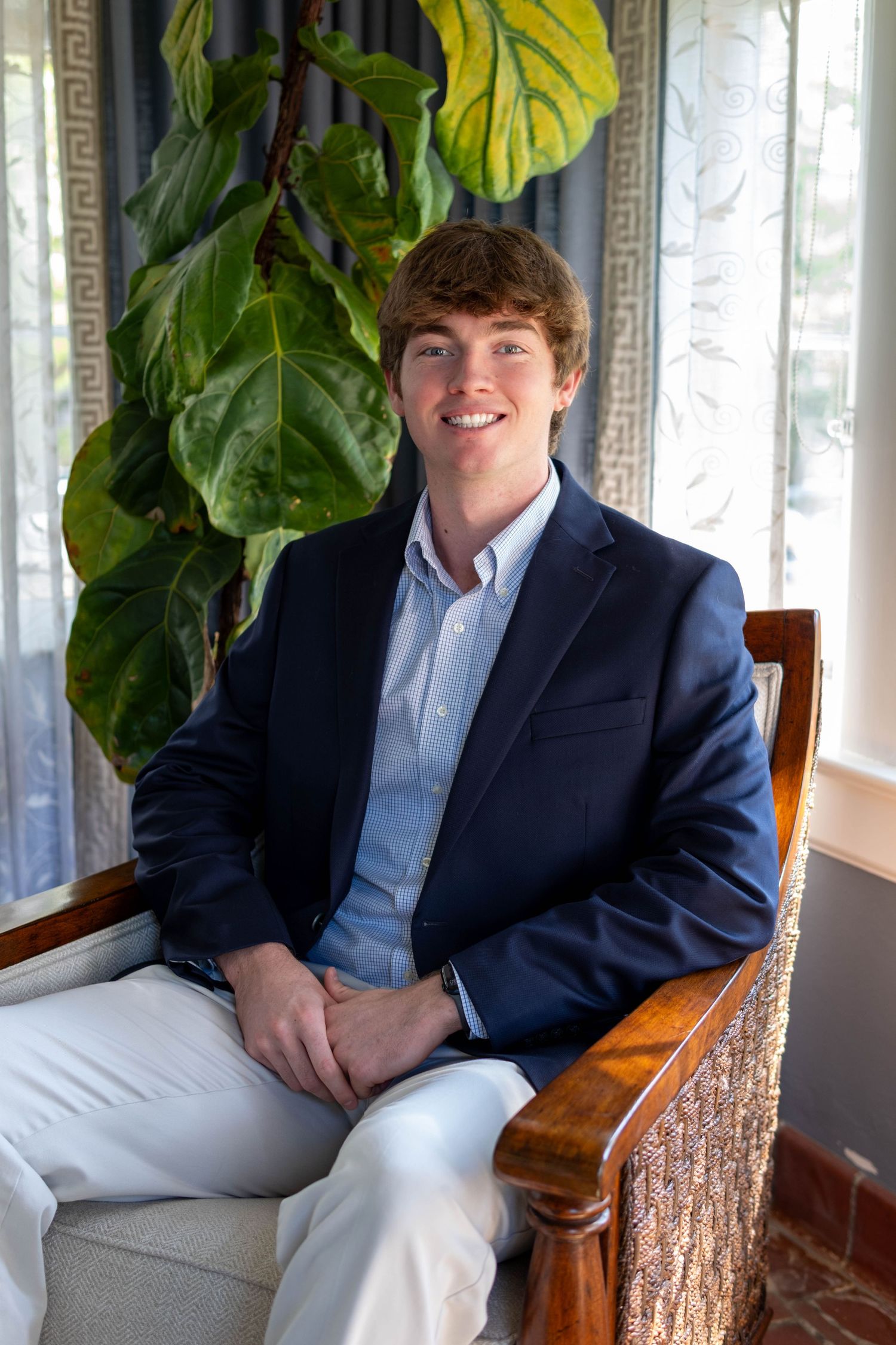 Young man in navy blazer sitting in wooden chair smiling with tropical plant background.