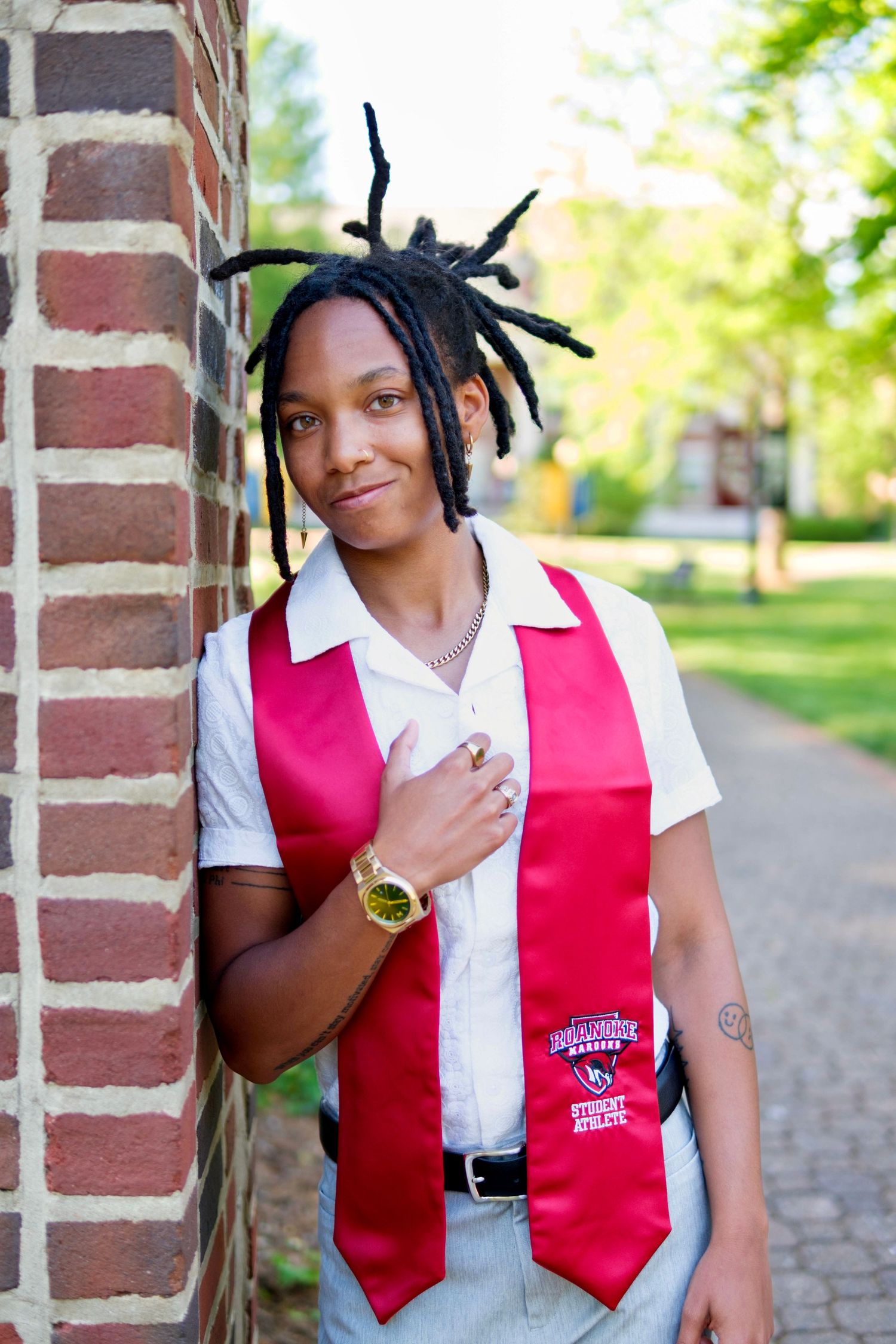 Graduate wearing red sash leaning against brick wall on sunny campus pathway.