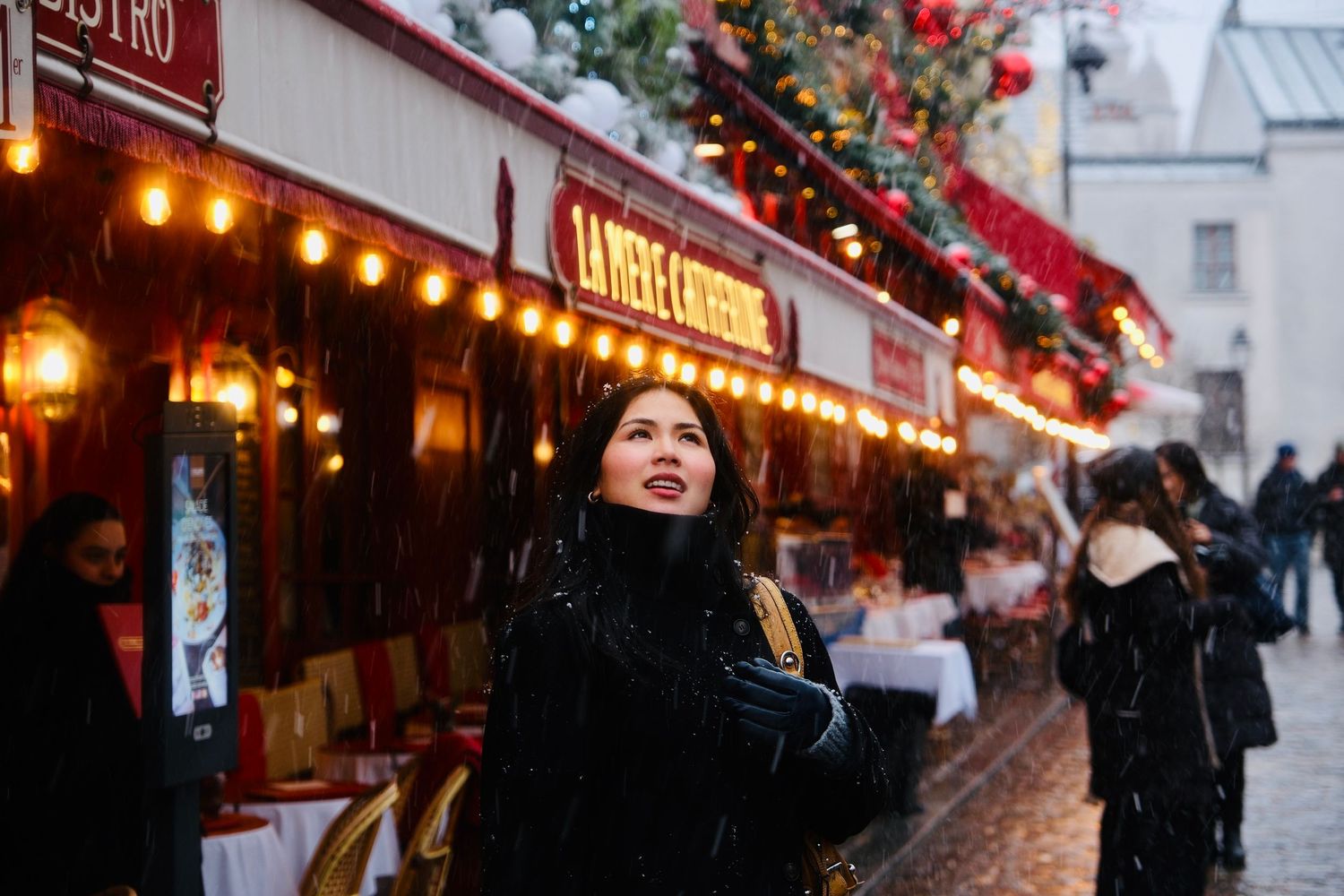 Young Asian woman standing outside a festively lit Parisian cafe during a snowy winter evening.