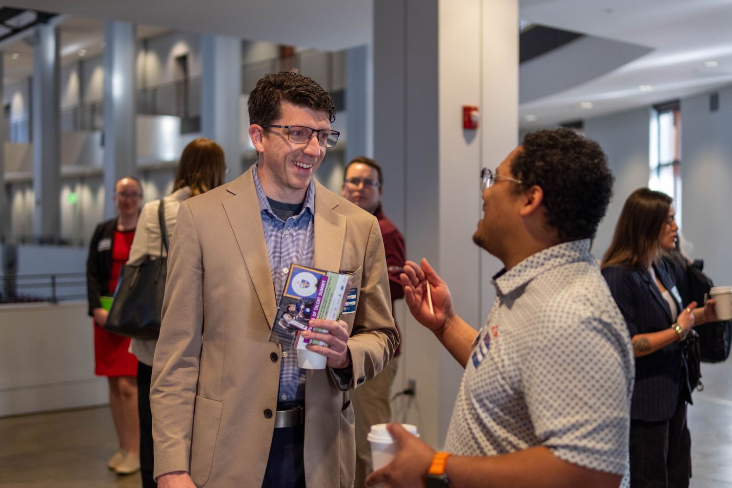 Two men in business casual attire smiling and conversing at indoor event.