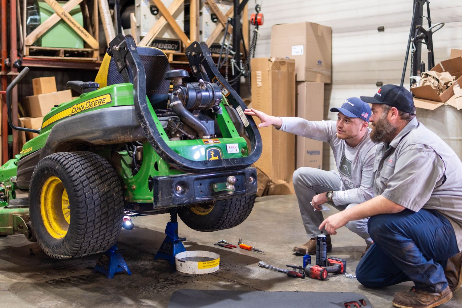 Two technicians repairing a John Deere riding lawn mower elevated on a lift in a workshop.