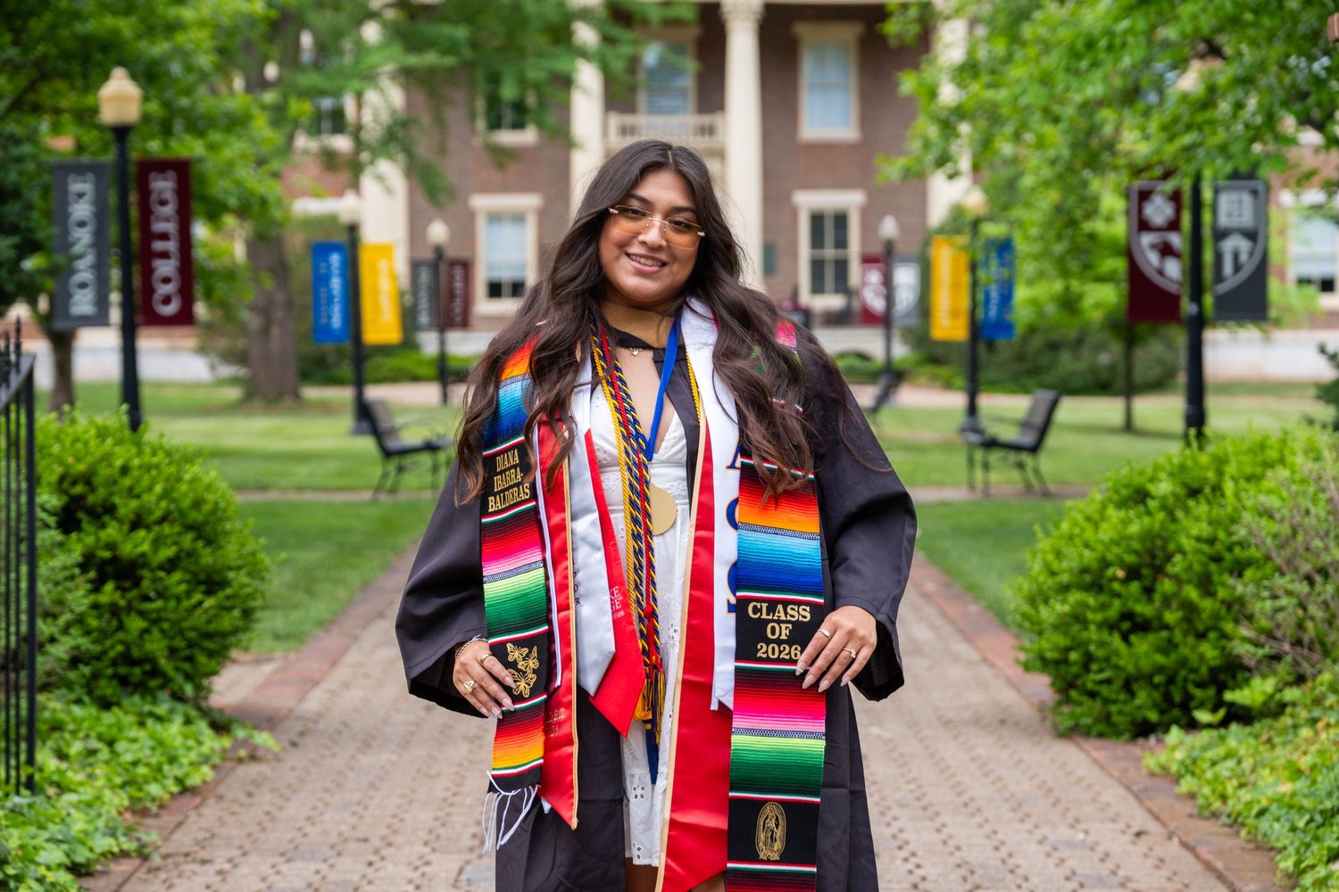 College graduate in cap and gown with colorful graduation stoles smiles on a brick campus walkway.