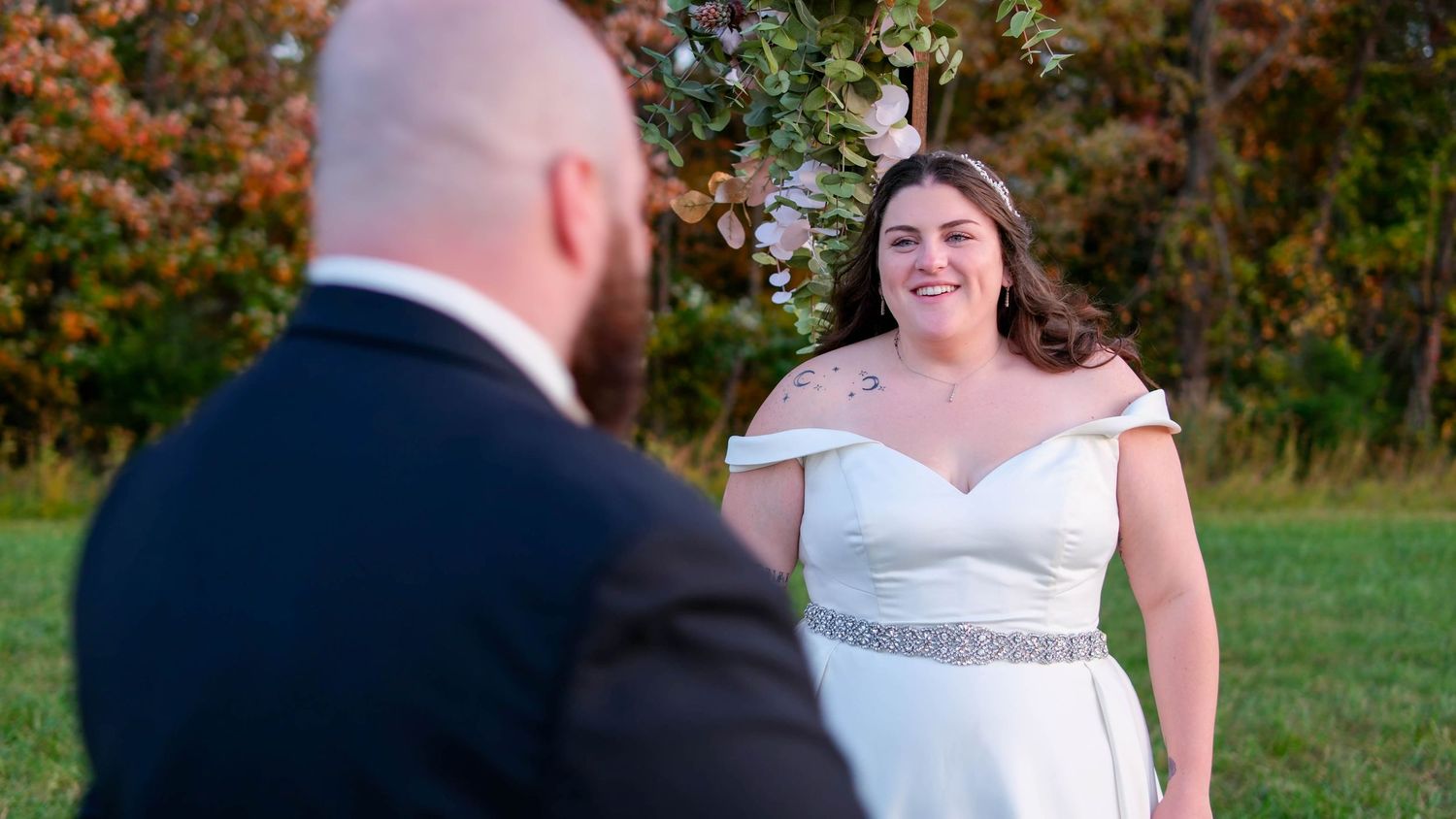 A smiling bride in an off-shoulder gown gazes at her groom during an outdoor fall wedding ceremony.