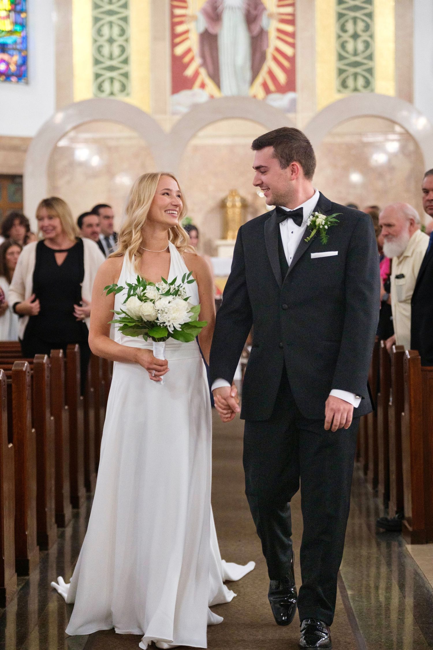 Bride and groom walk down church aisle after wedding ceremony, bride holding white floral bouquet.