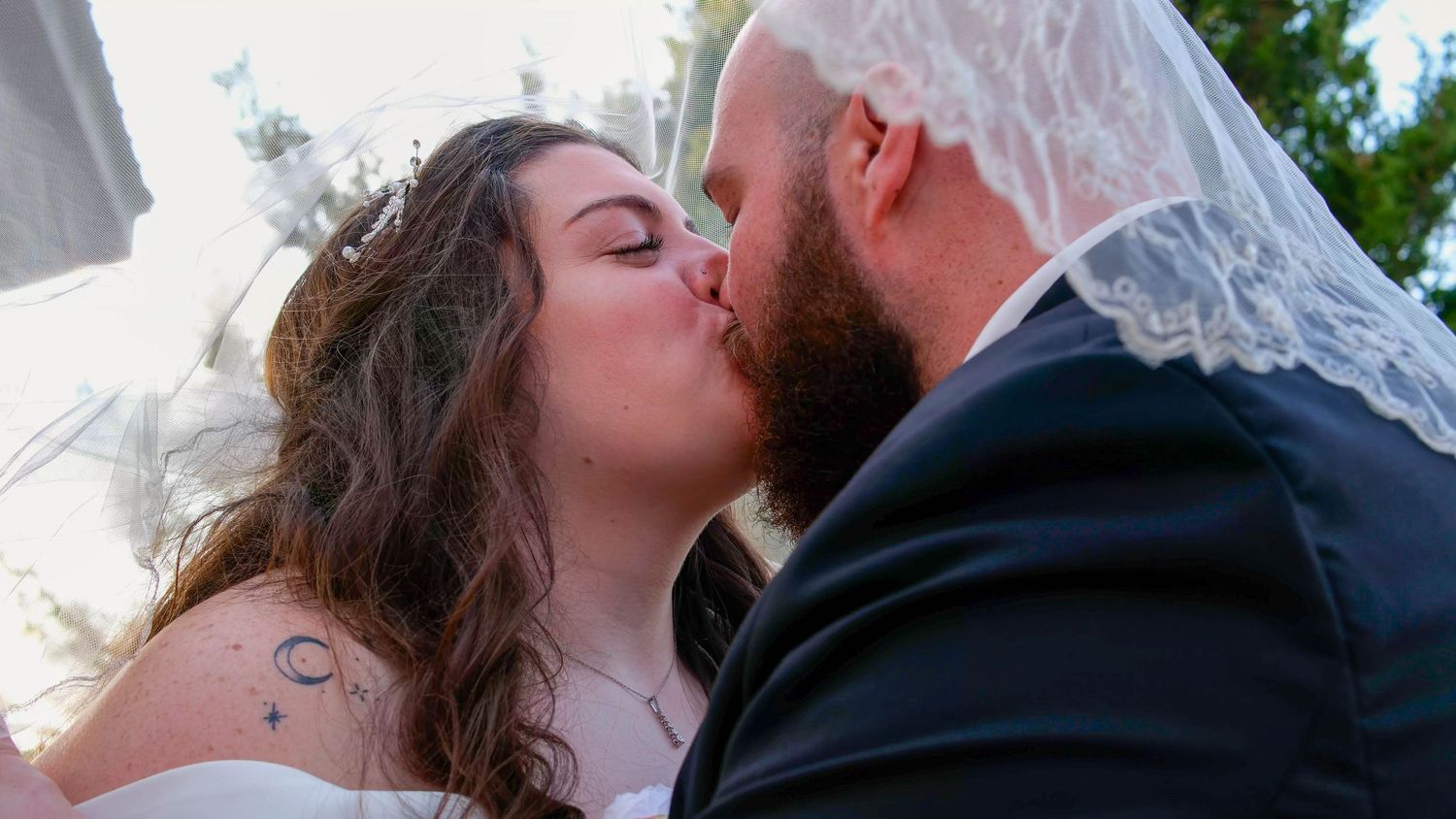 Newlyweds sharing a romantic kiss with bride in white dress and veil at outdoor wedding ceremony.
