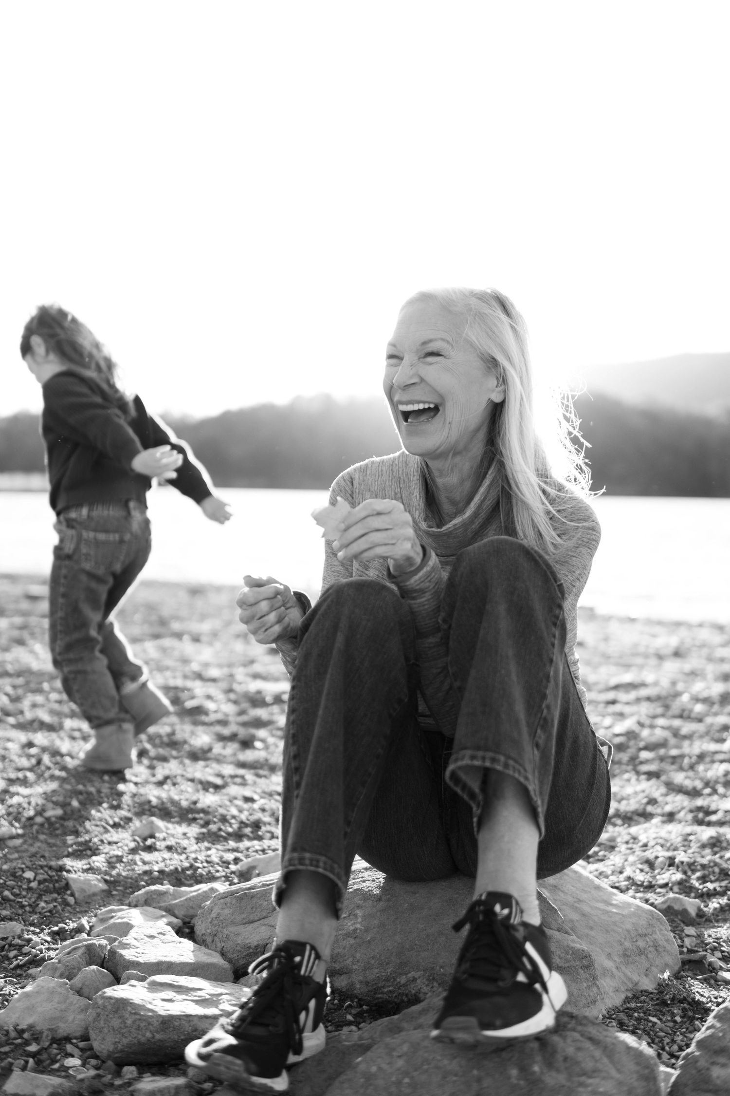 Black and white photo of a laughing blonde woman sitting on rocks outdoors near a lake.