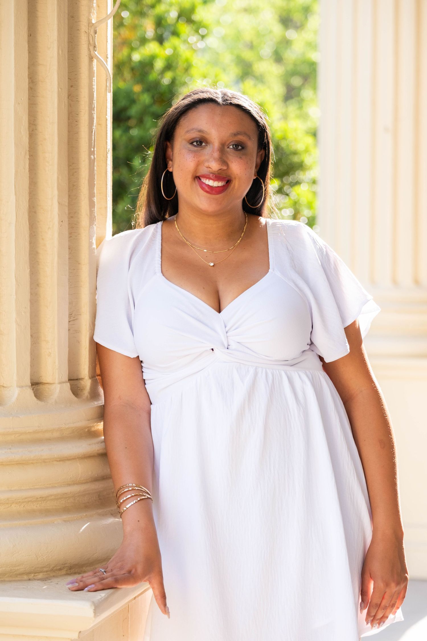 A smiling woman in a white dress with hoop earrings poses by classical columns outdoors.