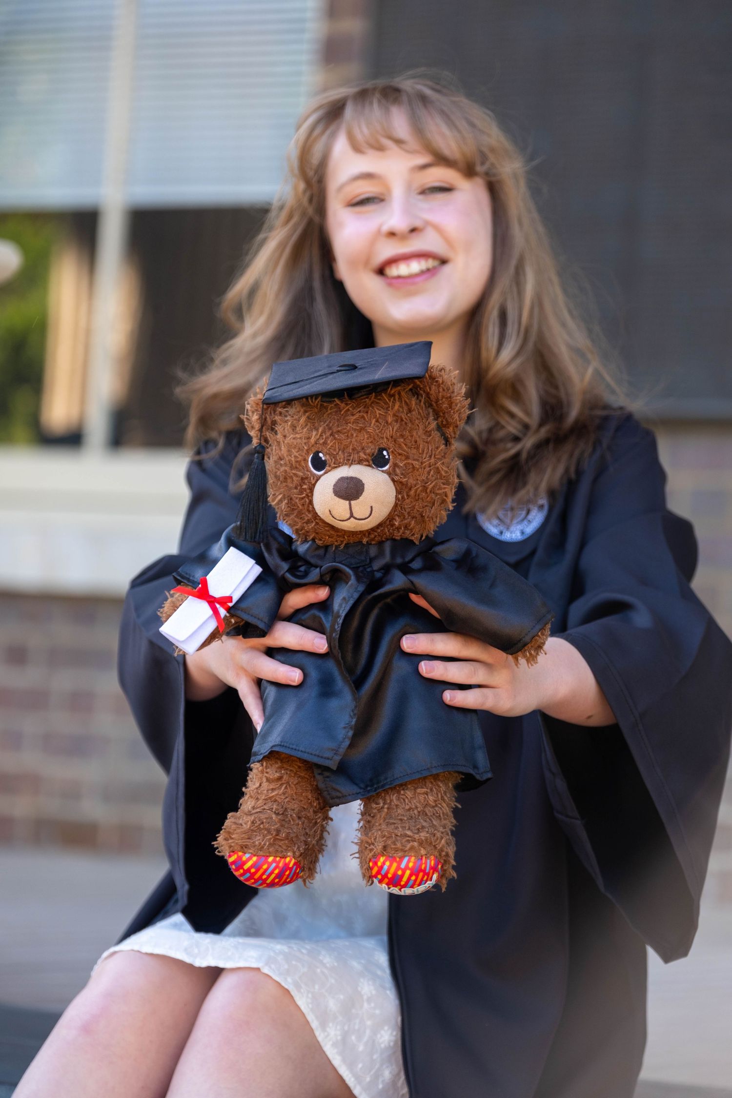 Smiling graduate in black gown holding a teddy bear dressed in matching graduation cap and gown with diploma.