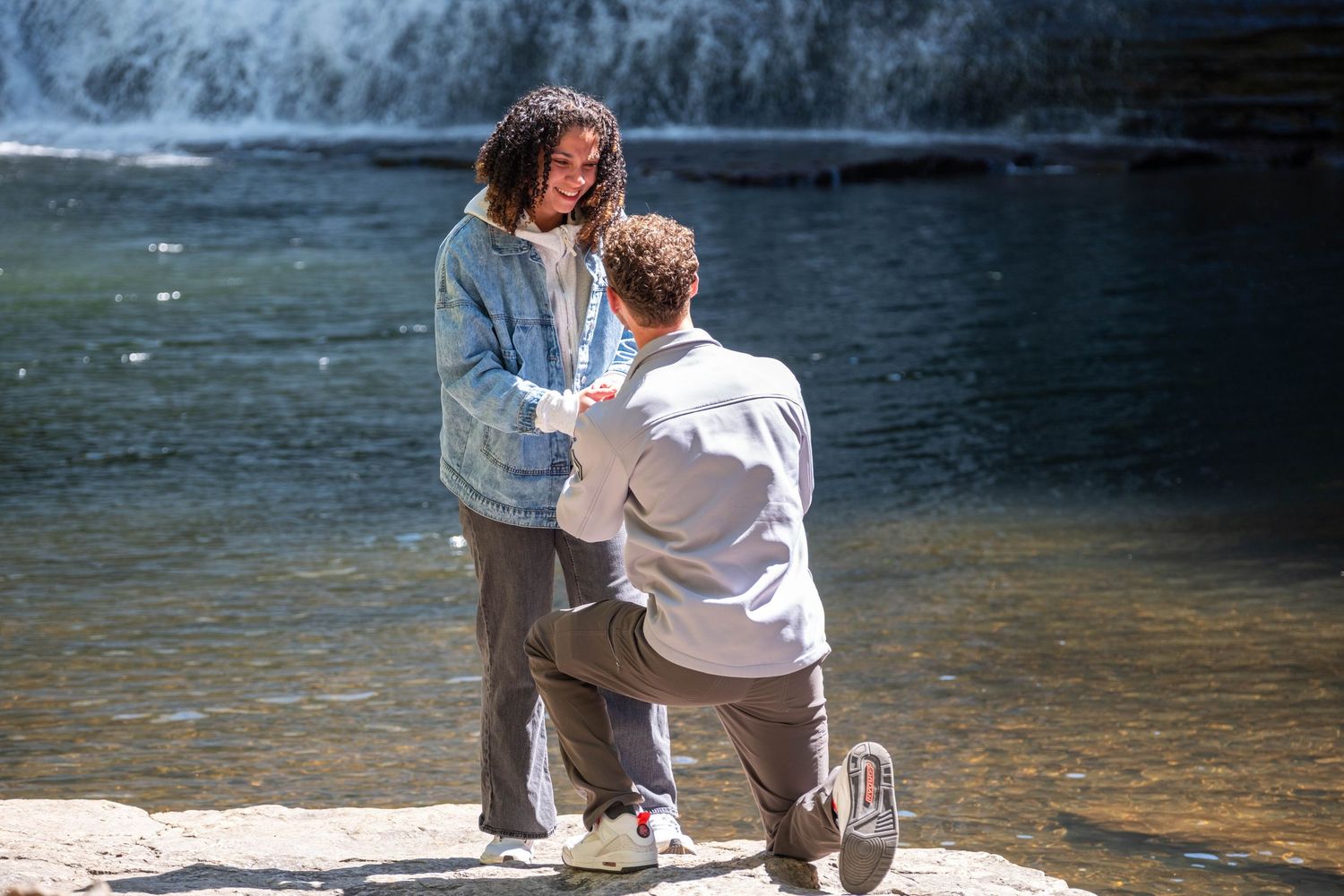 Man proposes on one knee to woman near a scenic waterfall on rocky shore in bright sunlight.