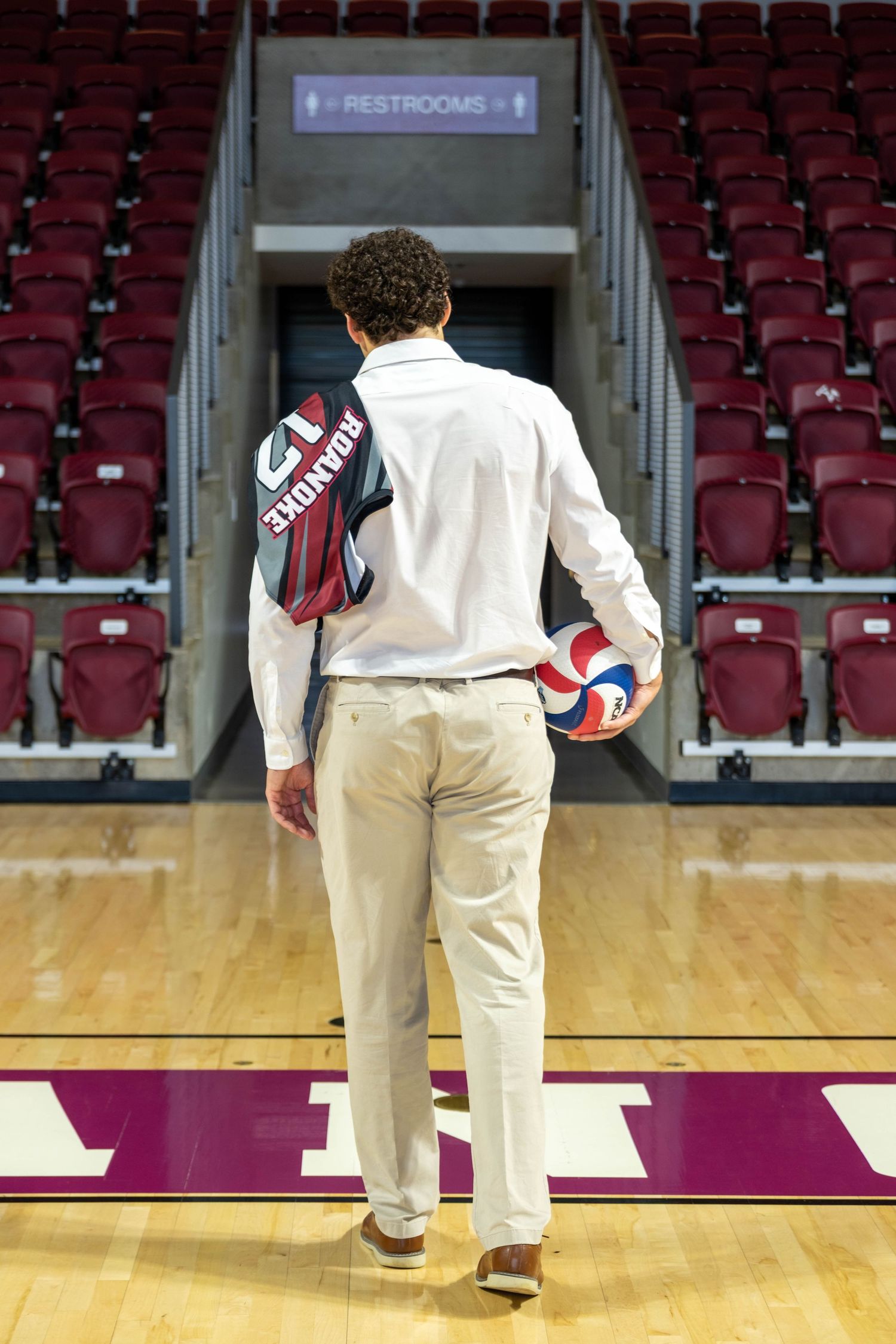 Young man in white shirt holding volleyball and sports bag walks toward arena seating on hardwood court.