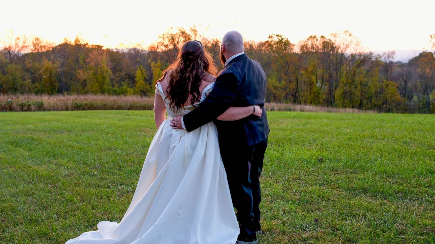 A bride and groom walk hand in hand across a green field at sunset with autumn trees in the background.
