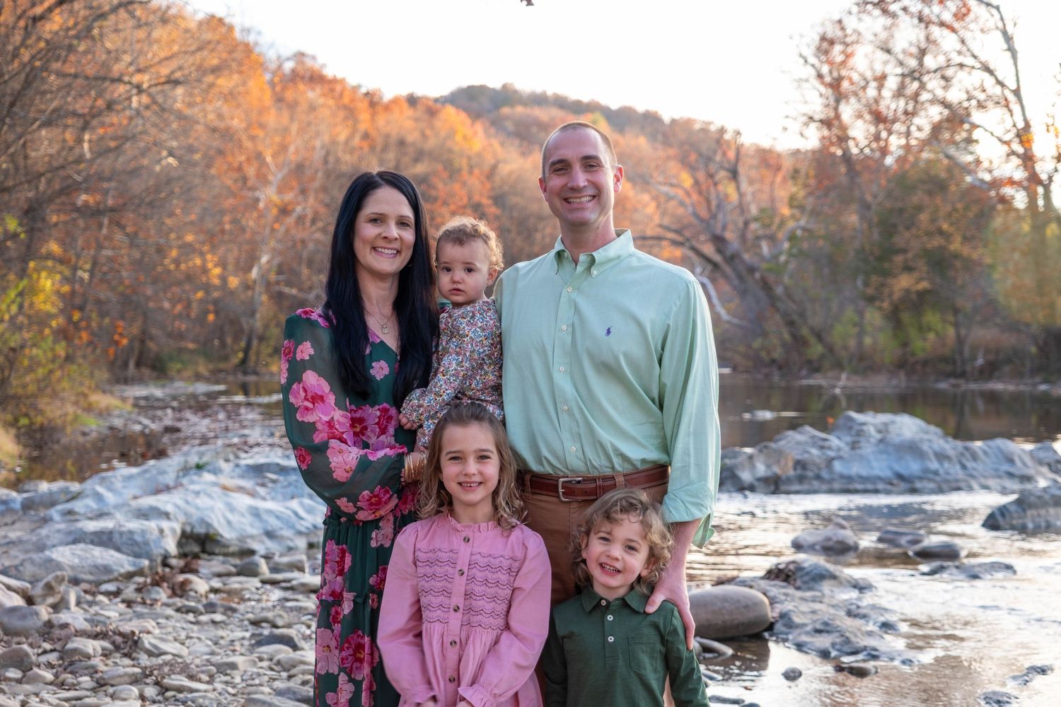 Family of five posing by a creek with colorful fall foliage in background.
