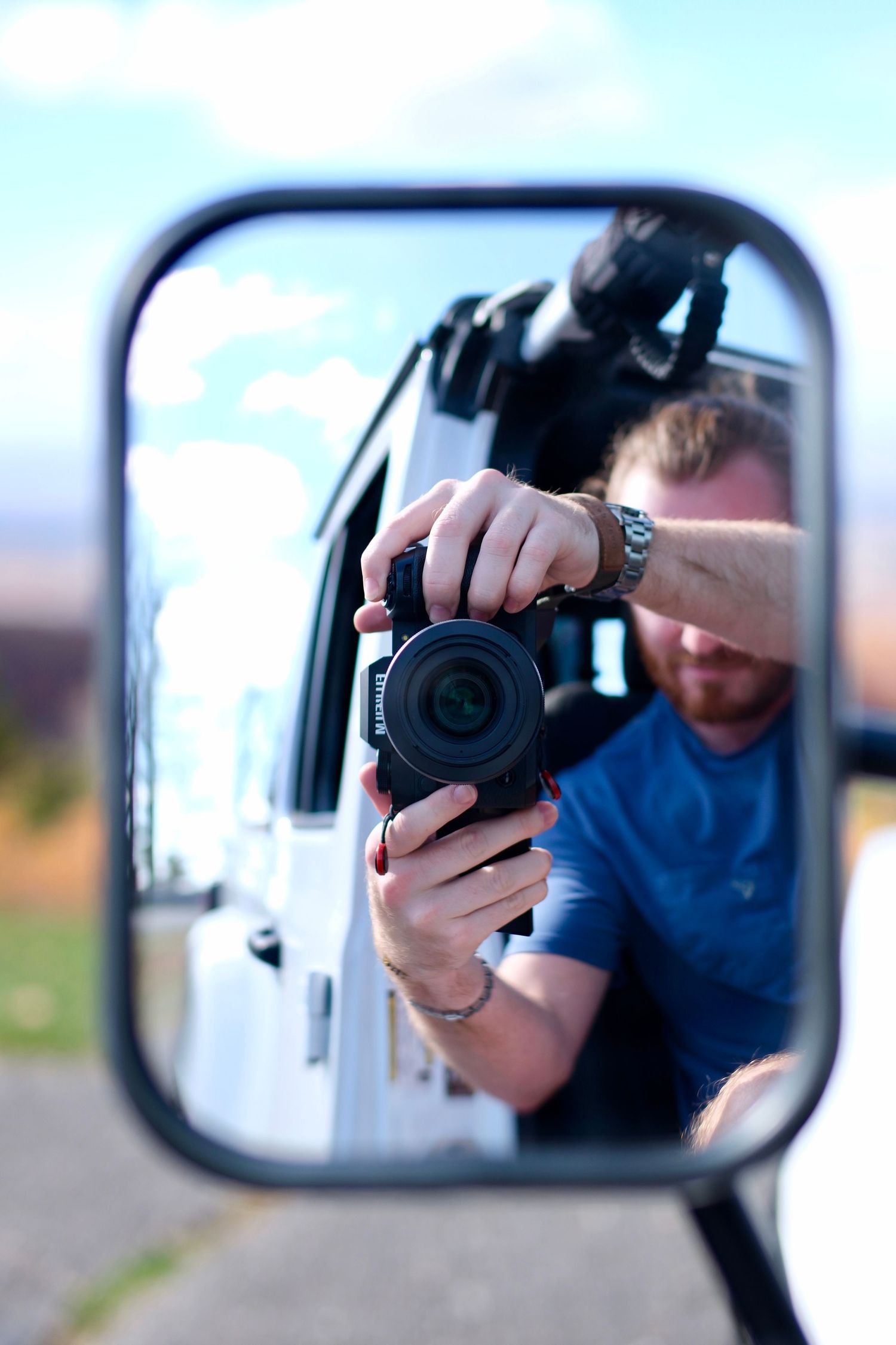 Bearded photographer capturing a self-portrait in a white van's side mirror outdoors.