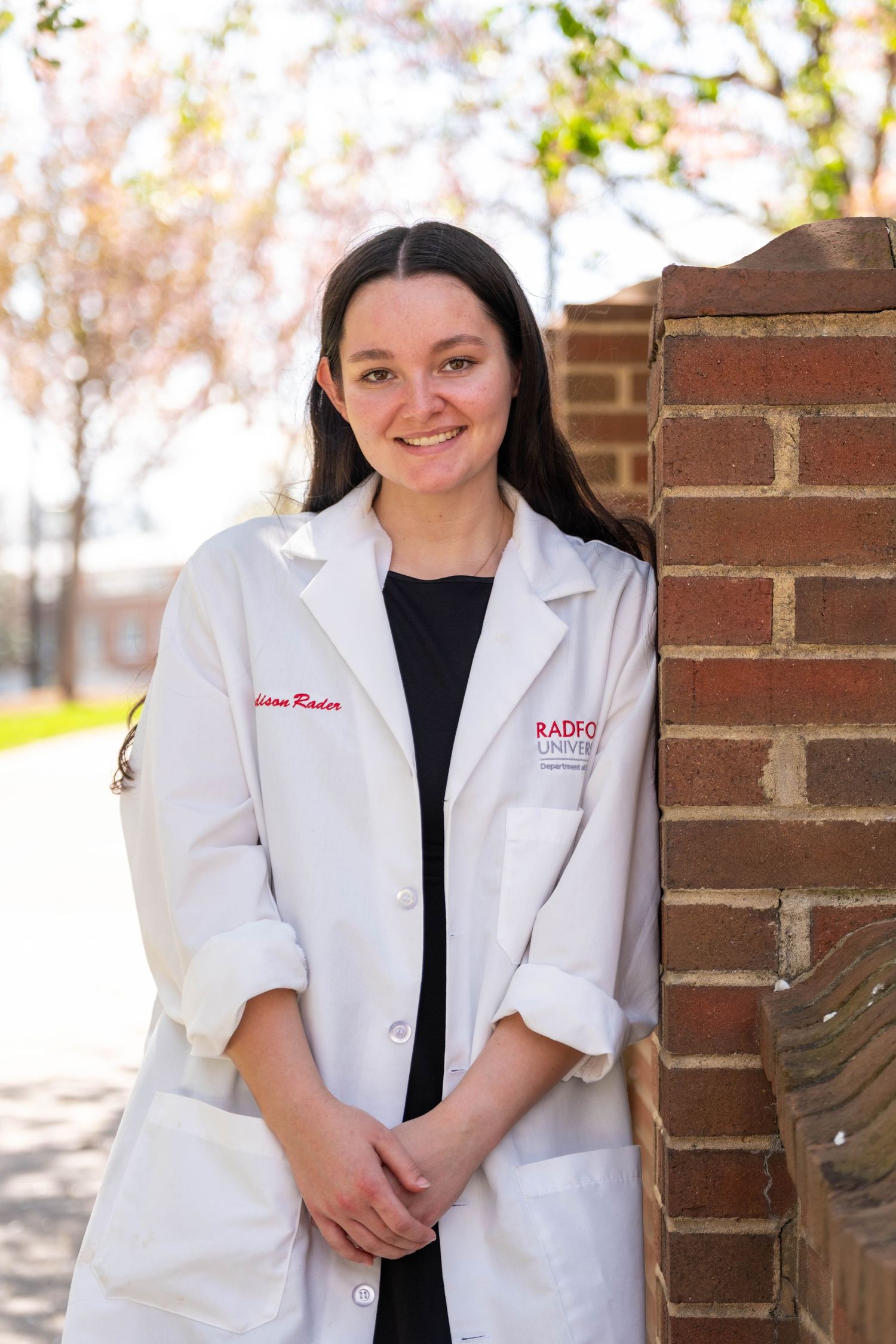 Smiling female student wearing Radford University white lab coat leaning against brick wall outside.