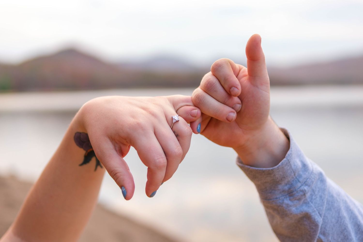 Close-up of couple holding pinky fingers near lakeside with small tattoo visible on womans wrist.