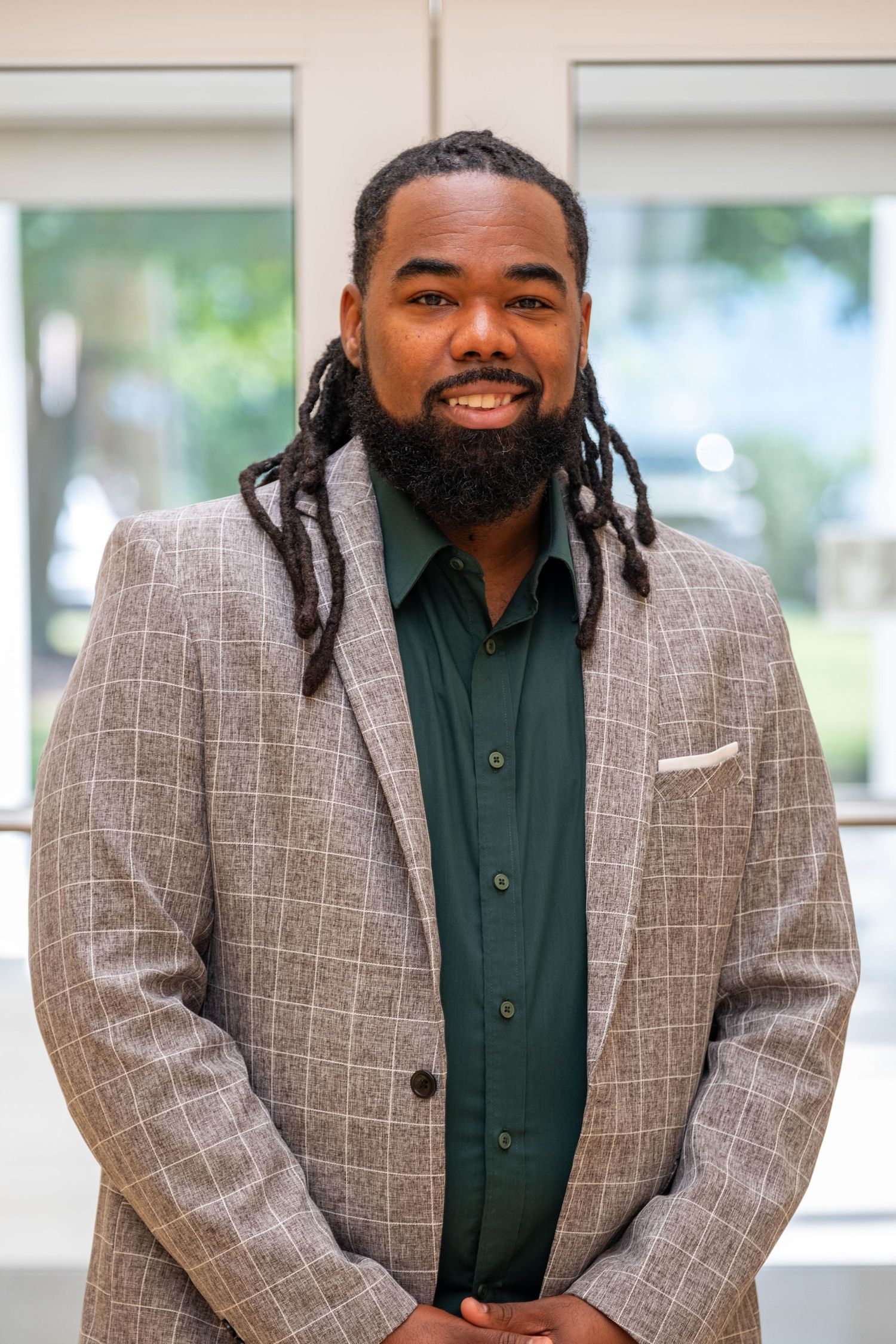 Professional man with dreadlocks and beard wearing a gray plaid blazer and green shirt, smiling indoors.