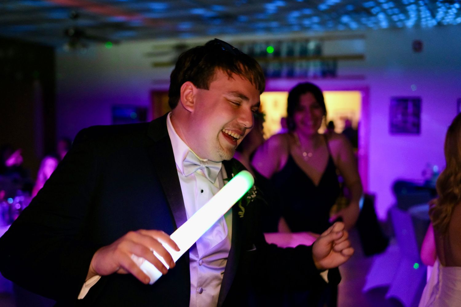 Excited man in tuxedo singing with glow stick on a colorful wedding reception dance floor.