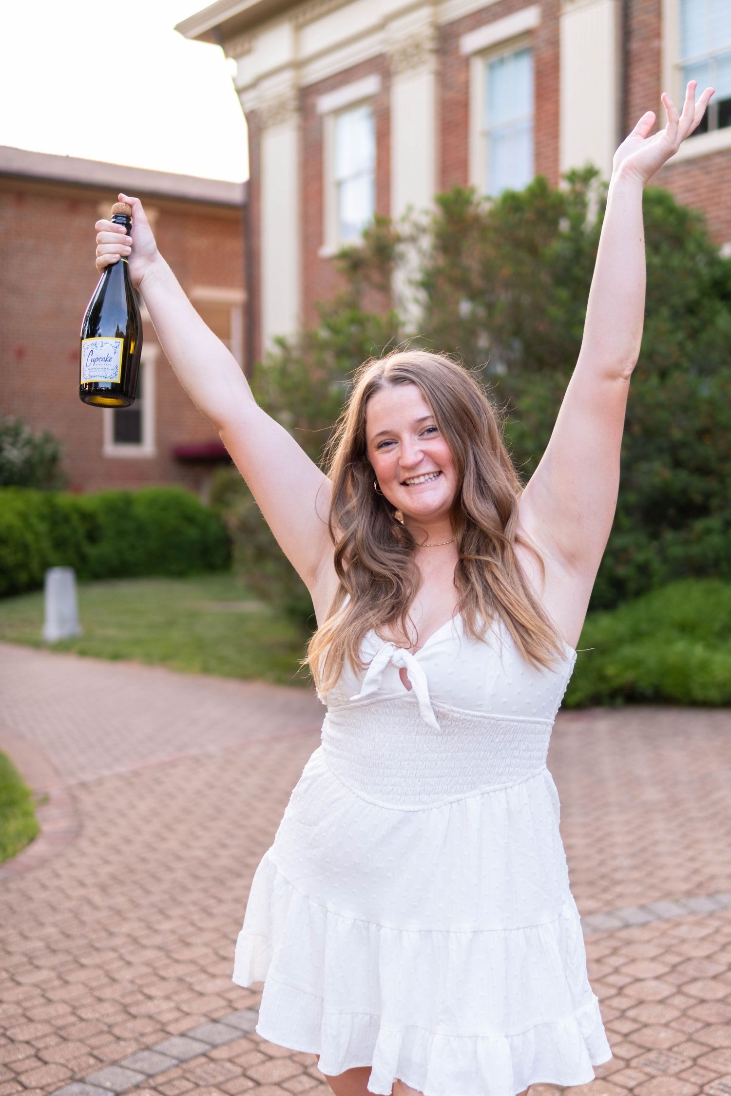 Joyful graduate in white dress celebrates with champagne bottle raised overhead on brick campus path.