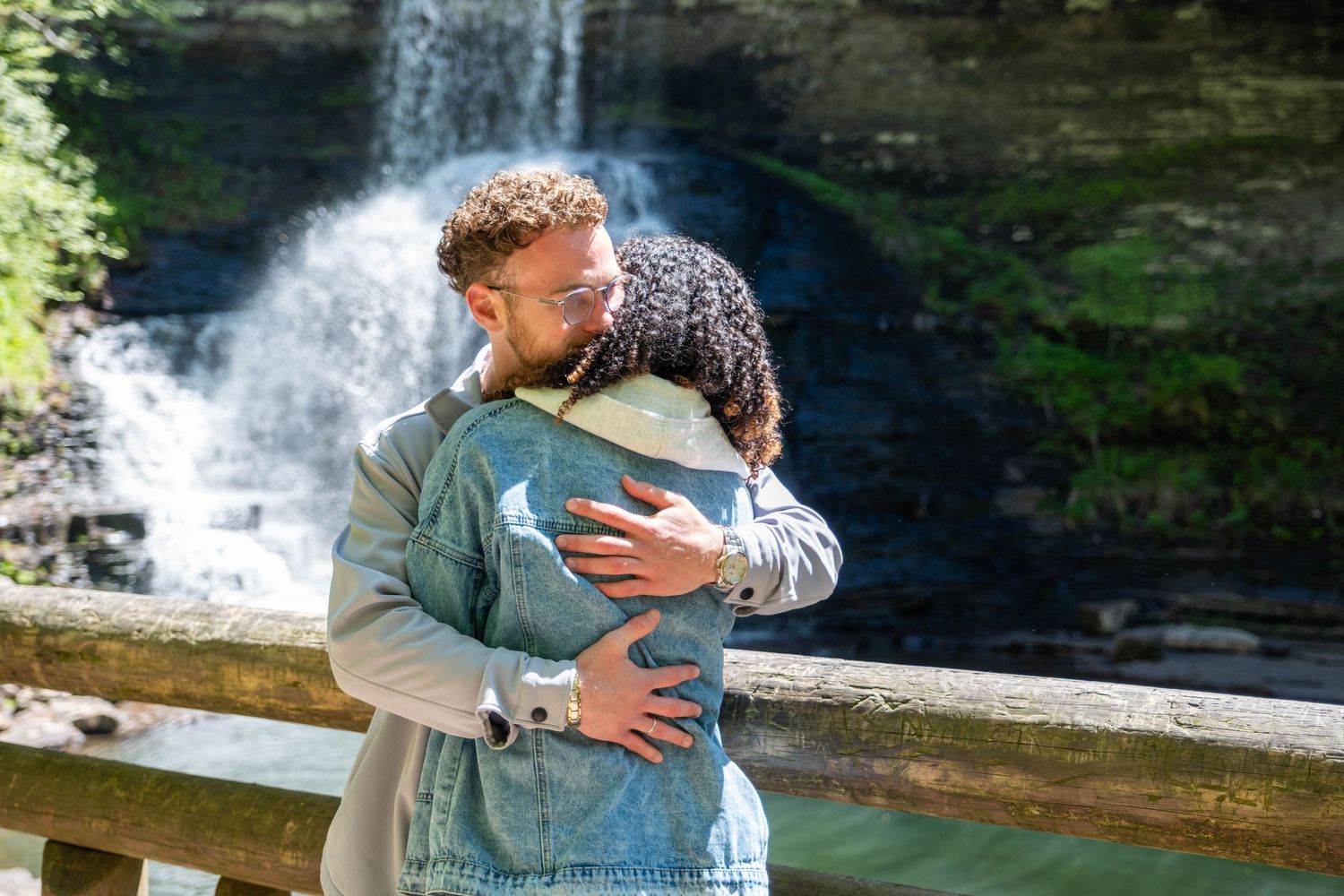 Couple embracing and kissing in front of a scenic waterfall on a sunny day near a wooden railing.